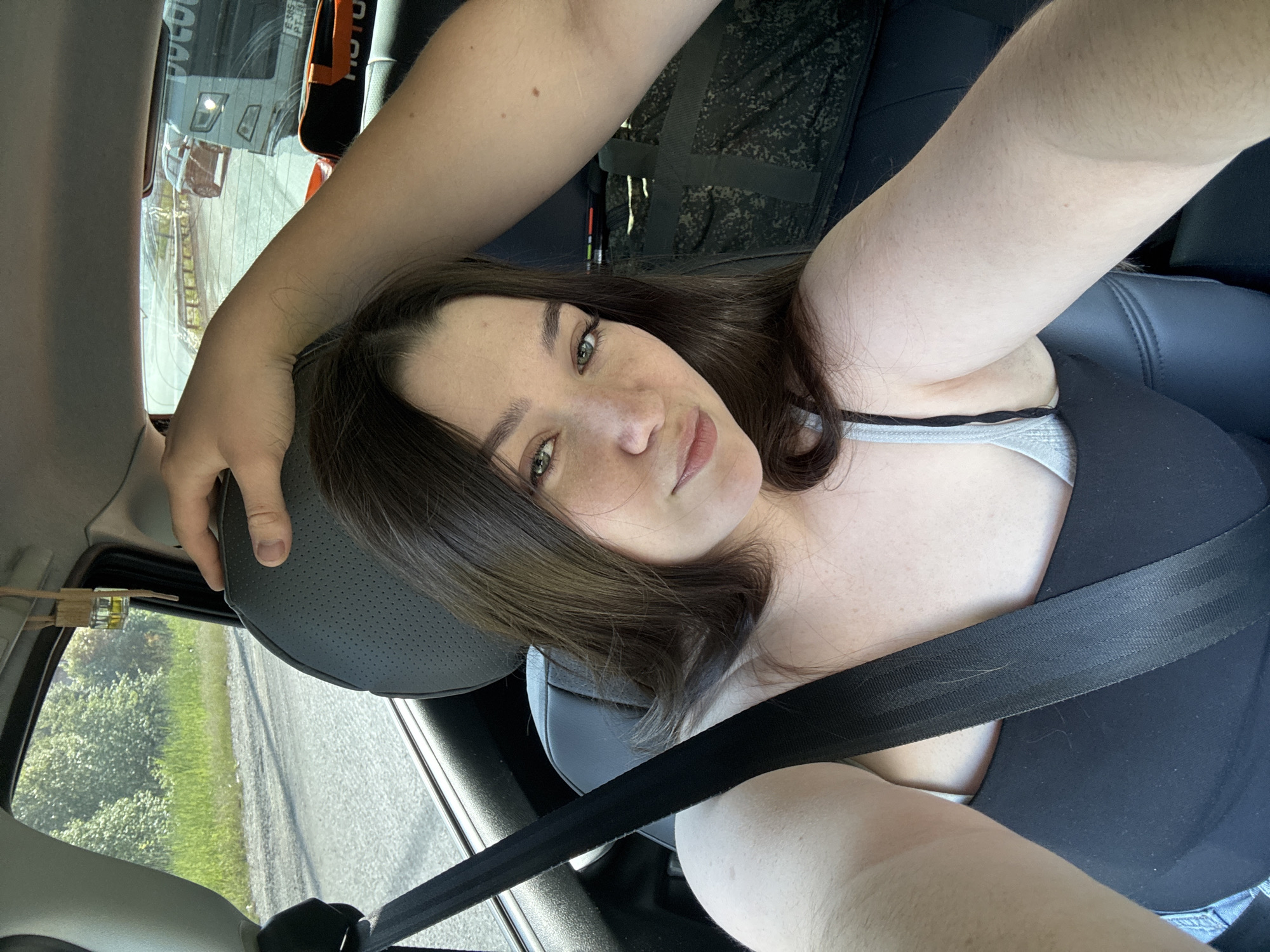 A woman with dark hair and green eyes is taking a selfie in the passenger seat of a car. She is wearing a black tank top and a grey bra. A seatbelt is across her chest. A hand is visible resting on the back of her headrest, suggesting another person is in the car. Through the car window, a road and trees are visible. In the background, a truck is driving on the road. The scene appears to be in Pesterevo, Russia.