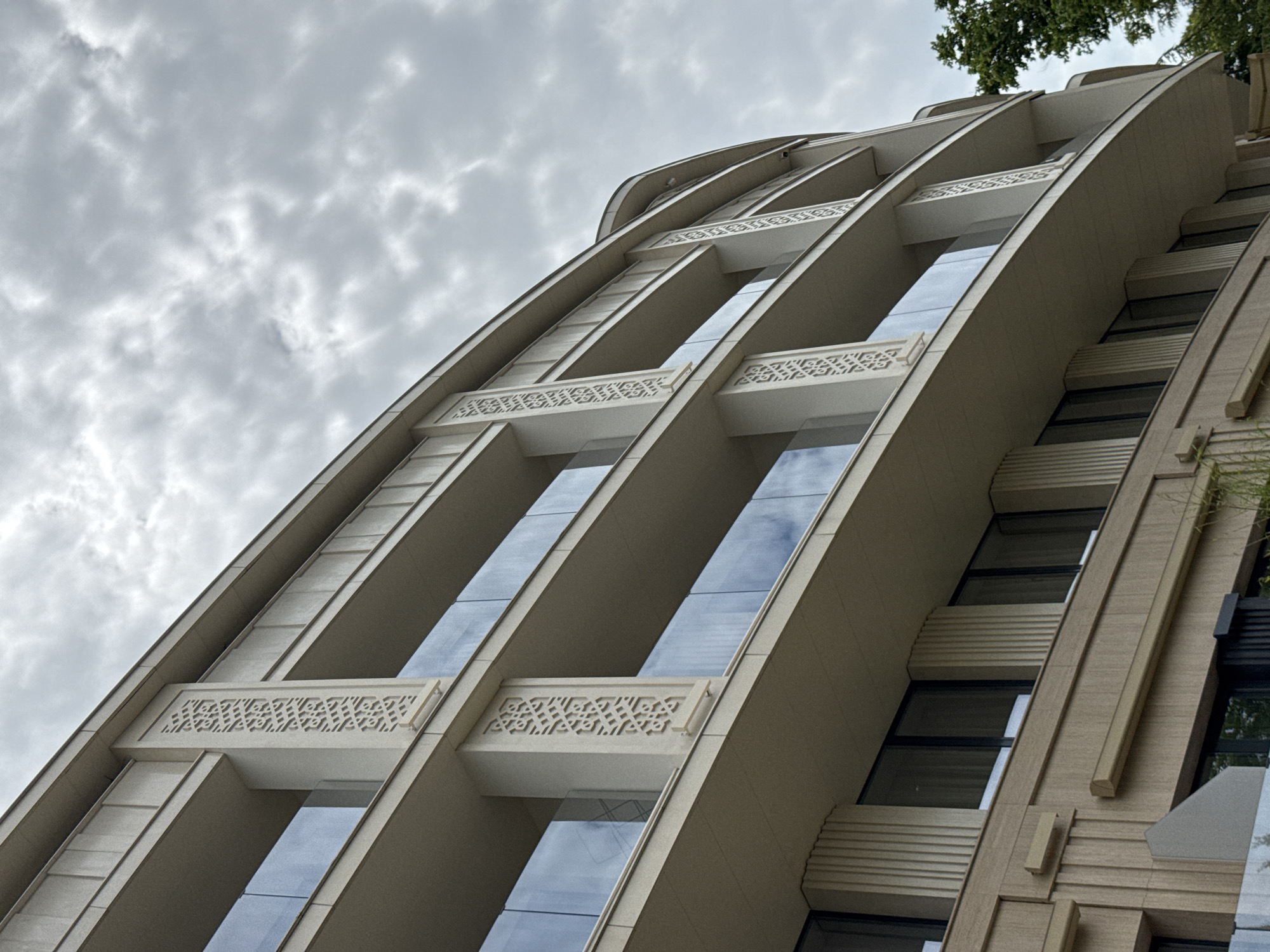 This is a low-angle shot of a modern building in Sochi, Russia, on a cloudy day. The building features multiple stories with balconies and glass railings. The facade is decorated with vertical fluting and geometric patterns on pillars between balconies. The sky is overcast with textured clouds. The lower part of the building shows windows and further decorative elements. A tree is visible on the right side of the frame.