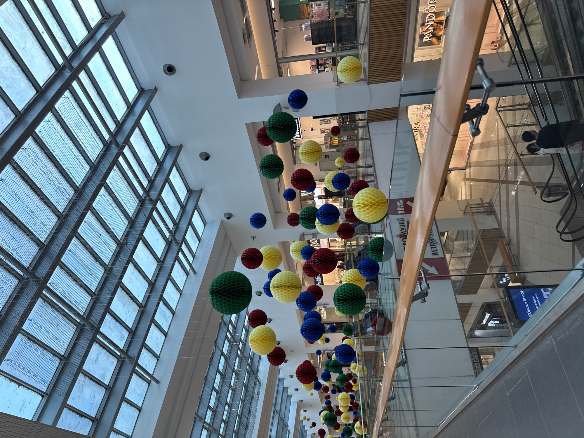 An indoor, multi-level shopping mall, observed from an upper floor looking down towards lower levels, is depicted in Unknown, Unknown. The ceiling features a prominent glass and metal-framed skylight, allowing natural light into the space, with a wire mesh structure visible beneath the glass panels. Numerous honeycomb paper spheres in green, yellow, red, and blue are suspended at varying heights throughout the mall's interior, creating a decorative arrangement.
The mall's architecture includes white walls, columns, and glass railings with light-colored wooden handrails that define the edges of each floor. Escalators connect the different levels. Storefronts are visible on multiple floors; notable signage includes "PANDORA" for a jewelry store and "РЕТРО МУЗЕЙ" (Retro Museum) on a lower level. A digital display screen by "JCDecaux Image" is also visible on a lower floor. Overhead, circular black security cameras are mounted on the white walls. A small sign near the bottom left indicates "1 floor этаж".
Two individuals are visible on an escalator ascending from the lowest floor: one person in a white hooded garment is to the left of another person wearing a dark top. They are moving downwards on the escalator. The overall environment appears well-lit and maintained, with a tiled floor visible on the upper level from which the photograph was taken.