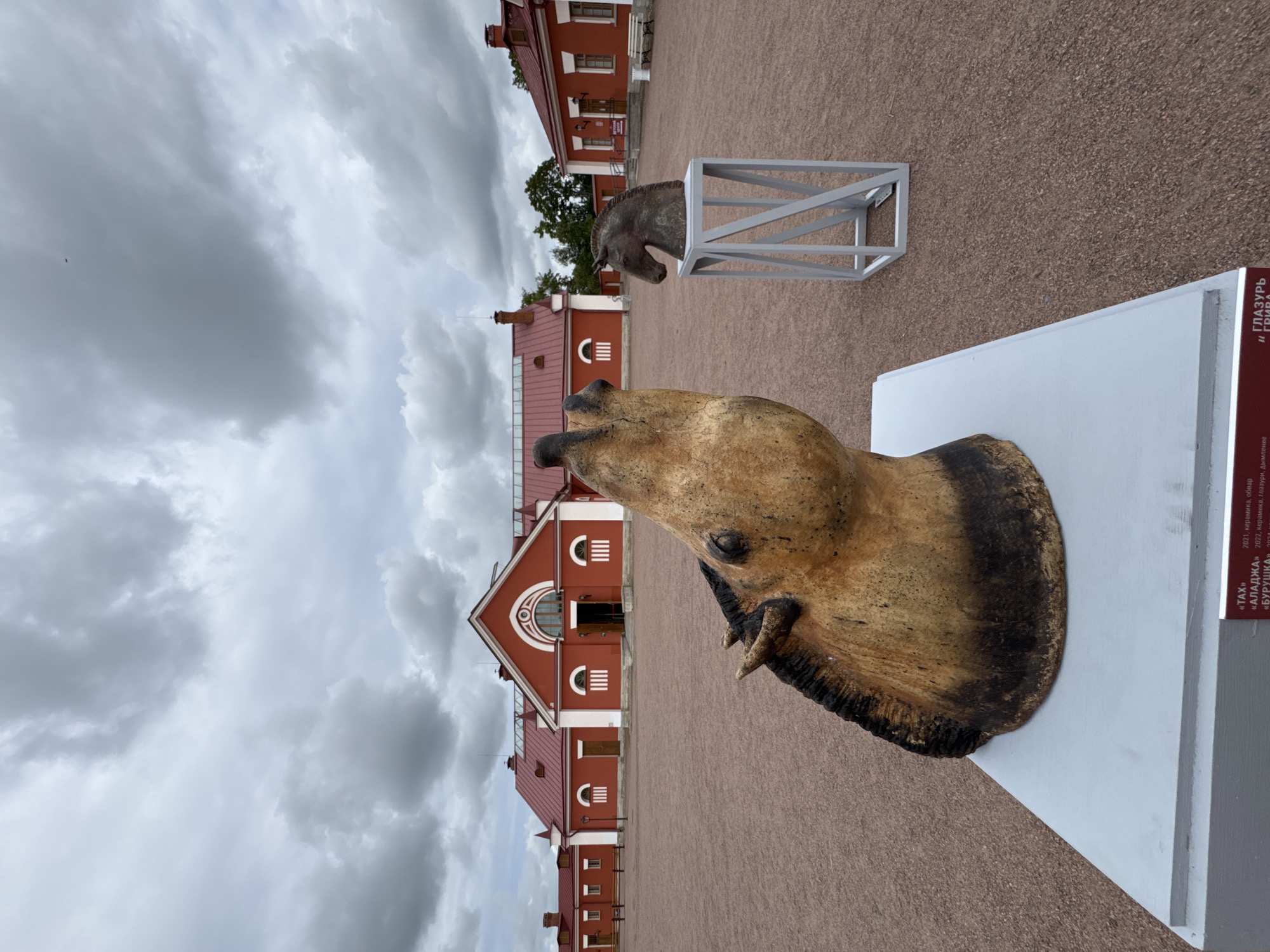 Two equestrian sculptures are displayed in an outdoor courtyard. The sculpture in the foreground is a large, tan and black ceramic horse head, tilted upwards, on a white pedestal. Behind it and to the right, a smaller, dark bronze-colored horse head sculpture sits on a modern, geometric gray stand. In the background, a row of red buildings with white trim and red roofs, likely part of the Pavlovsk Palace ensemble in Pushkin, Russia, stretches across the frame. The courtyard is paved with reddish-brown gravel. The sky is overcast with gray clouds.