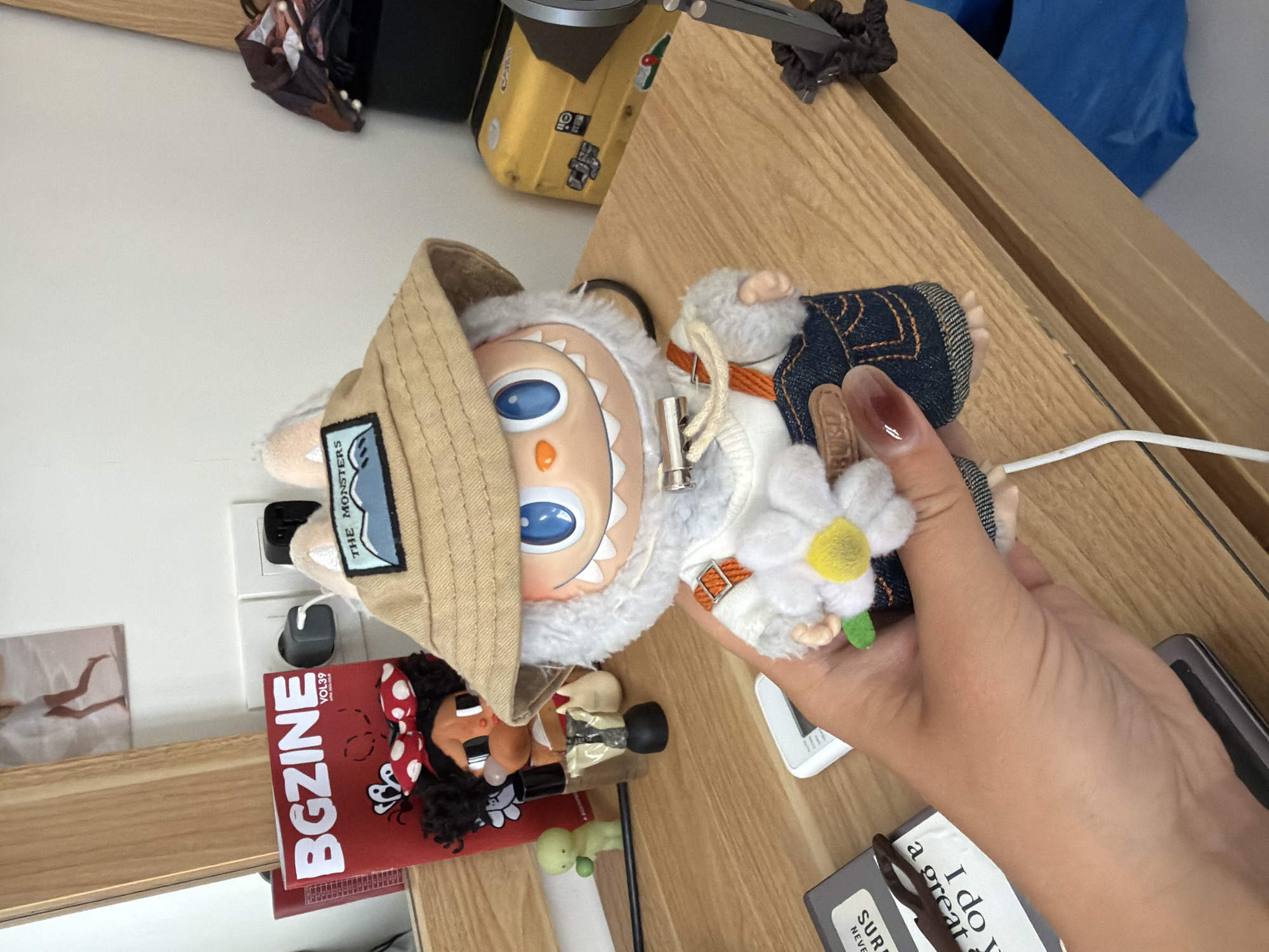 The image shows a cluttered tabletop in what appears to be an indoor setting, possibly a residential space. Dominating the view is a furry toy figure with a tan hat, being held by a hand. The hand, with visible nail polish, grasps the toy near a white daisy-like flower accessory attached to it.
Around the toy, various objects are scattered on the table. There is a red magazine with the word "BGZINE" visible, a small doll figure with black hair and a red and white polka dot bow, a green object, a grey case with glasses, a white electrical adapter plugged into a wall socket, a small photograph, and a yellow object with black details. Parts of a wooden shelf or table frame are also visible.
The location, Kuta Selatan, Indonesia, does not appear to be visually evident from the contents of the image alone. No recognizable landmarks, cultural artifacts, or specific environmental features of Kuta Selatan are present.