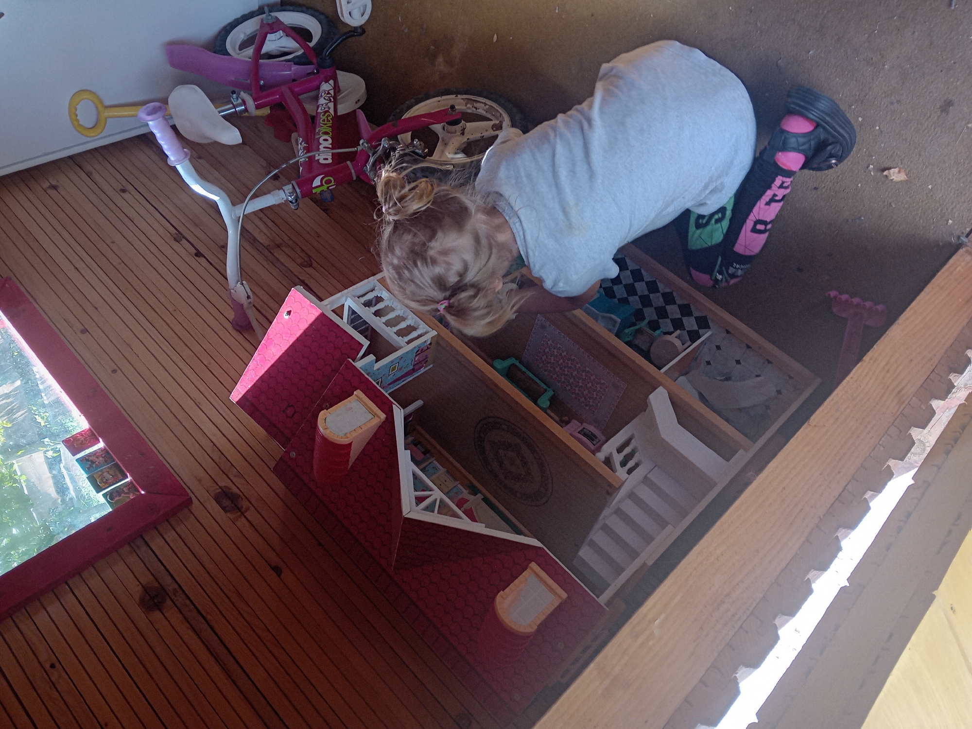 The image depicts a young girl in a wooden structure, possibly a playhouse or shed, in Zuberec, Slovakia. The girl, with blonde hair in pigtails, is wearing a grey t-shirt and is kneeling on the floor. She appears to be interacting with a dollhouse, which is positioned to her left. The dollhouse is red and white with multiple levels and windows. To the right of the dollhouse is a pink children's bicycle with training wheels and a yellow handle. The floor is a mix of wood planks and what appears to be linoleum. A window with visible greenery outside is present in the upper left corner of the structure.