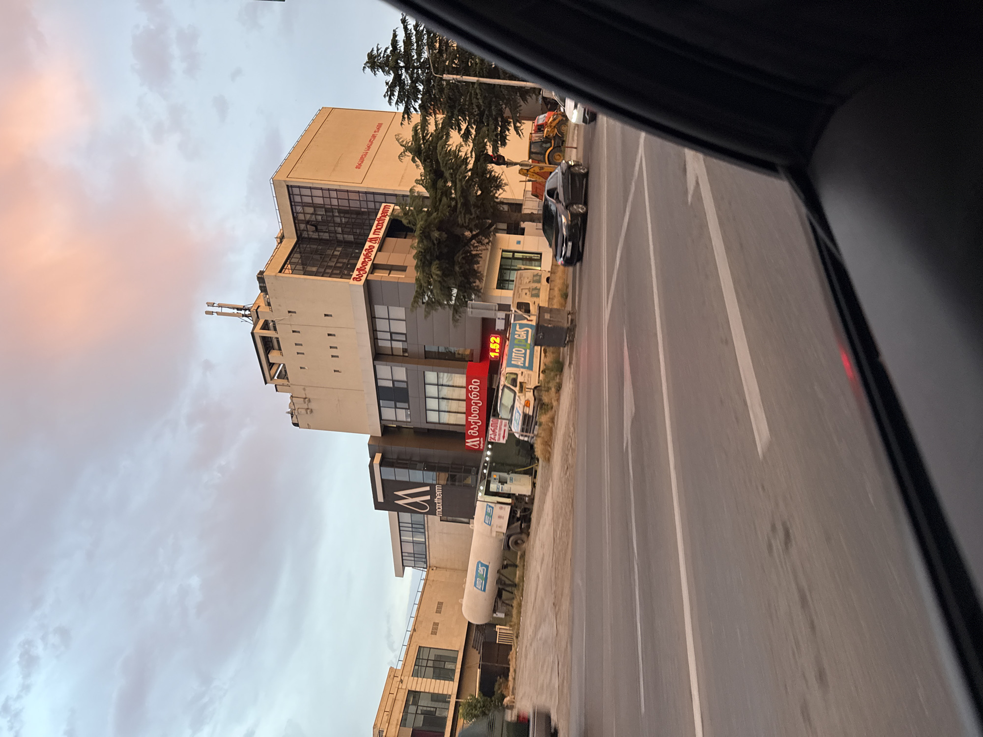 The image depicts a street scene in Tbilisi, Georgia, likely captured from inside a vehicle. The time of day appears to be late afternoon or early evening, with soft, warm light from the setting sun illuminating the sky with hues of pink and orange, contrasting with the cooler gray clouds.
The primary subjects are a multi-story building and a street with vehicles. The building is modern in design, with a beige facade on its upper floors and darker glass and metal on the lower levels. Prominently displayed on the building are signs with text in Georgian. One red sign reads "მაესტრო," and another, a vertical red sign with white text, says "auto gas." Below this, a sign with a white "W" logo and the text "maxtherm" is visible.
Several vehicles are present on the street. A black car is driving on the right side of the road, and a white tanker truck, possibly for fuel, is parked on the side. In the background, a yellow construction vehicle, likely a backhoe loader, is also visible. Trees, including a tall evergreen, line the street, providing some greenery against the urban backdrop.
The overall impression is of a typical urban environment during a transitional time of day, with everyday activities like driving and the presence of businesses. The sky suggests a pleasant, calm weather condition. The orientation of the image is portrait, suggesting it was taken with the camera held vertically.