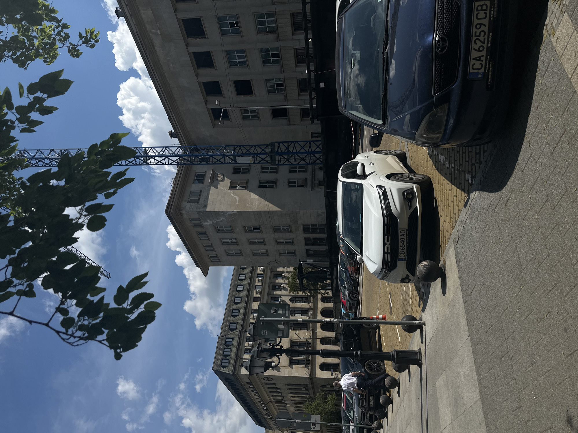 The image captures a street scene in Sofia, Bulgaria. A line of parked cars occupies the right side of the frame, with a blue Toyota Corolla (license plate CA 6259 CB) and a white Dacia Duster (license plate CB 6540 AO) prominently displayed. A sidewalk runs adjacent to the cars, featuring bollards and a lamppost. A man is seated next to a parked car.
The background showcases tall, aging buildings with numerous windows. In the upper left, tree branches and leaves partially obscure the view of a cloudy sky. A construction crane is visible between the buildings. The buildings' architecture and condition suggest a central or historical area of Sofia.
