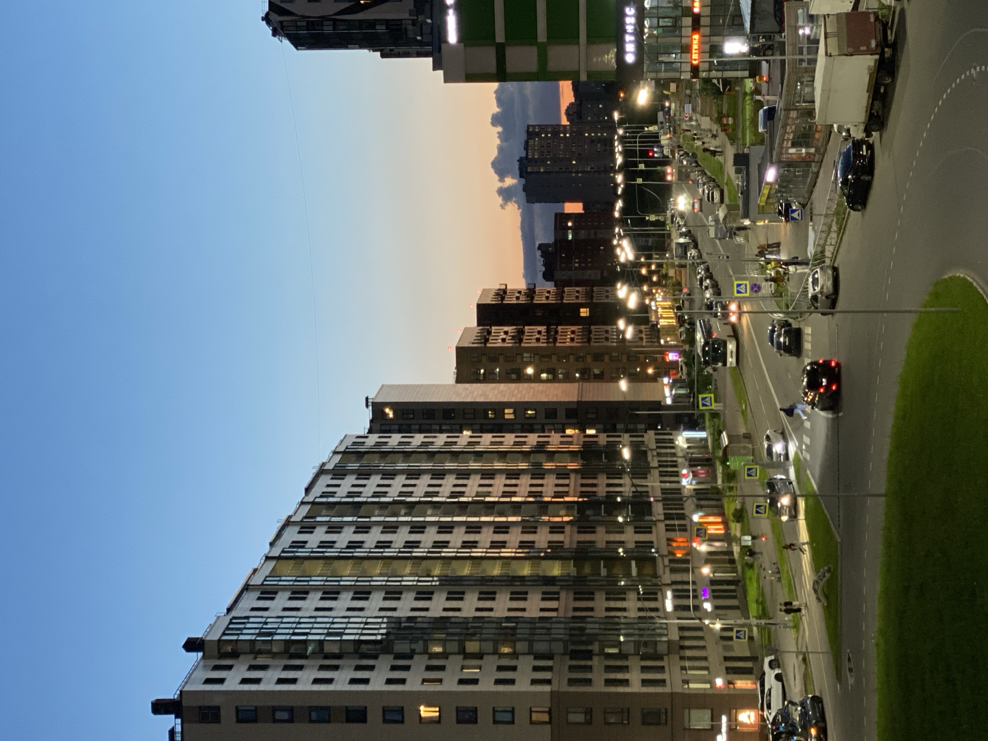 This image depicts a street scene in P.im.sverdlova, Russia, during the twilight hours. The sky transitions from a clear blue overhead to warmer hues near the horizon, with some clouds present. Dominating the left side of the frame are several tall, modern apartment buildings, characterized by their uniform window patterns and light-colored facades. Reflections of the sky and the sunset are visible in the windows of these buildings.
A multi-lane road runs through the center of the image, with cars and a bus in motion. Streetlights are illuminated along the road, casting a glow on the asphalt and surrounding areas. Pedestrians are visible on the sidewalks and crossing the street at designated crosswalks. Several smaller commercial buildings with illuminated signage are situated along the right side of the road. The overall impression is one of an active urban environment transitioning into evening.