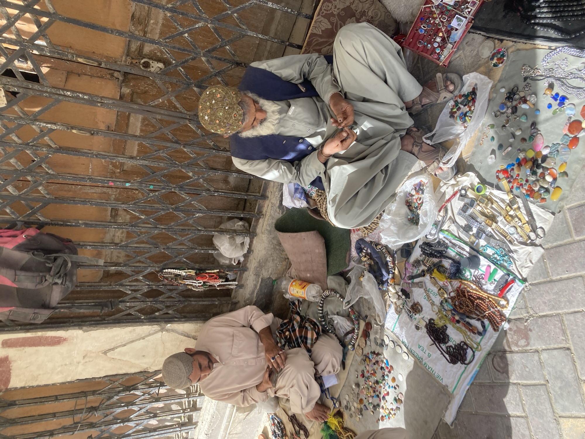 Two men sit on the ground selling jewelry and other items in Quetta City Tehsil, Pakistan. The older man is wearing a patterned cap and a grey outfit with a blue vest. The other man is wearing a light tan outfit and a grey cap. The goods are spread out on the ground in front of them. The items include jewelry, beads, stones, watches, and other small objects. A metal gate is in the foreground.