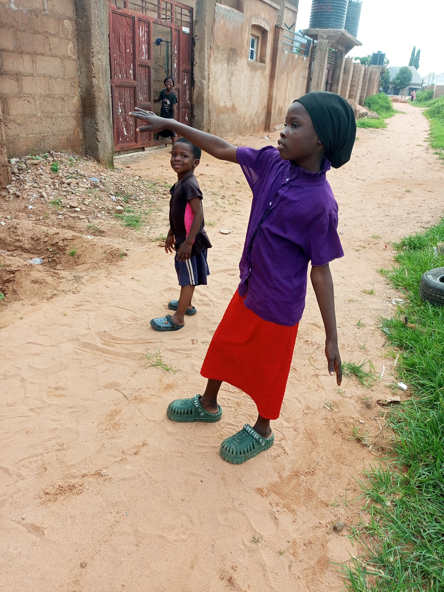 In Tempure, Nigeria, a young child wearing a black headscarf and a purple shirt over a red skirt is captured mid-motion on a sandy dirt road. The child's right arm is extended outwards, palm facing up, as if reaching for or pointing to something beyond the frame. Their left arm hangs loosely by their side, with the hand dangling downwards. The child is wearing green perforated sandals on both feet. To the left of the main subject, another child, dressed in a dark t-shirt with pink accents and navy shorts, smiles at the camera. This child is also wearing blue perforated sandals. In the background, a building with a large, weathered, reddish-brown double door stands against a brick wall. A third, less distinct figure can be seen through the doorway. The environment consists of a sandy path bordered by sparse vegetation and a building structure in the distance, with large water tanks visible on top of one of the structures. The overall scene suggests an outdoor, everyday moment in a Nigerian community.