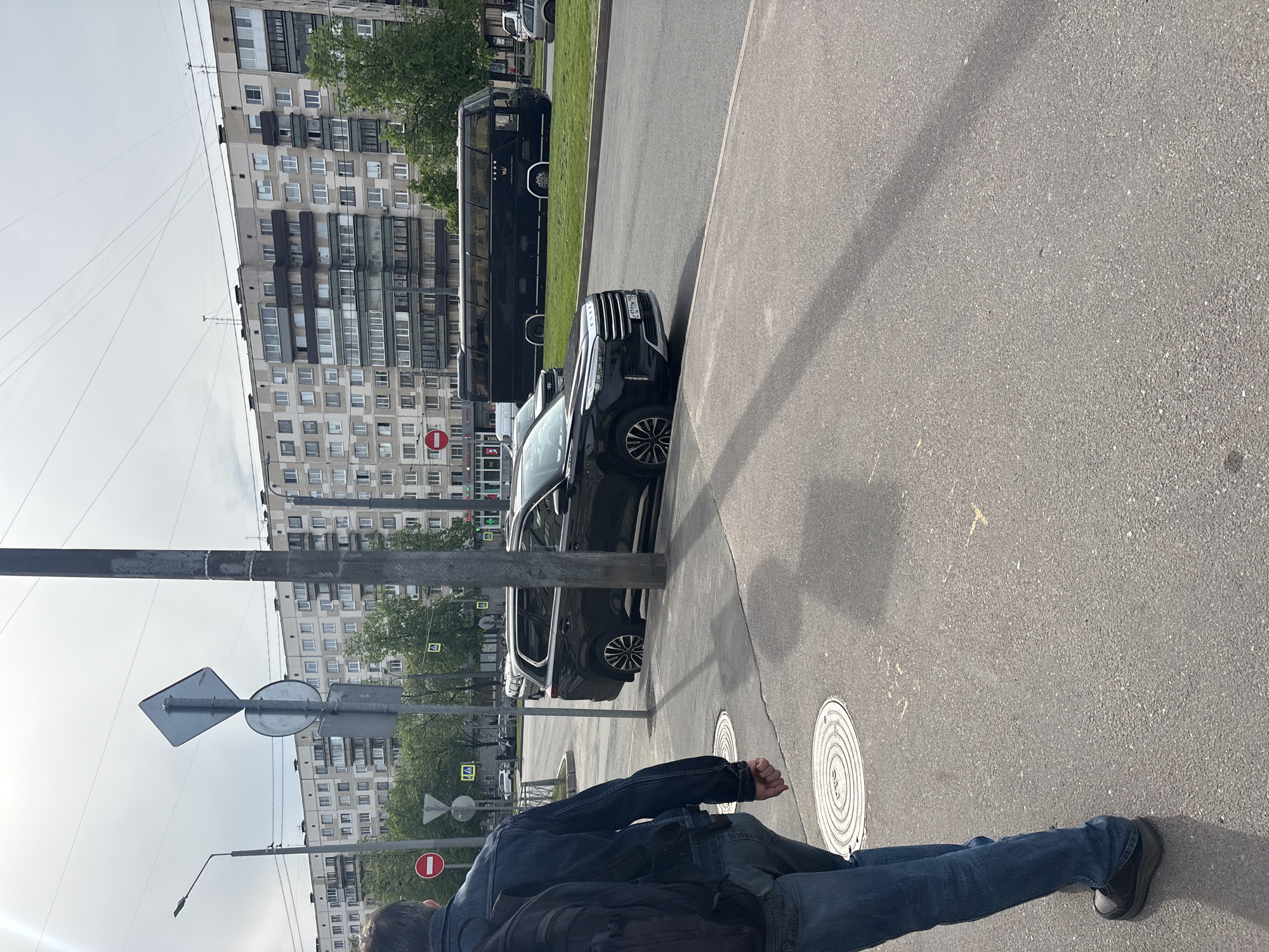 The image depicts an urban street scene in Saint Petersburg, Russia, with a building facade of approximately ten stories in the background. Parked along the roadside are a black passenger vehicle and a black tour bus. A person wearing a blue jean jacket, dark backpack, and blue jeans is walking along the pavement adjacent to the road. Street signs and a metal light pole are in the foreground, along with a manhole cover on the pavement.