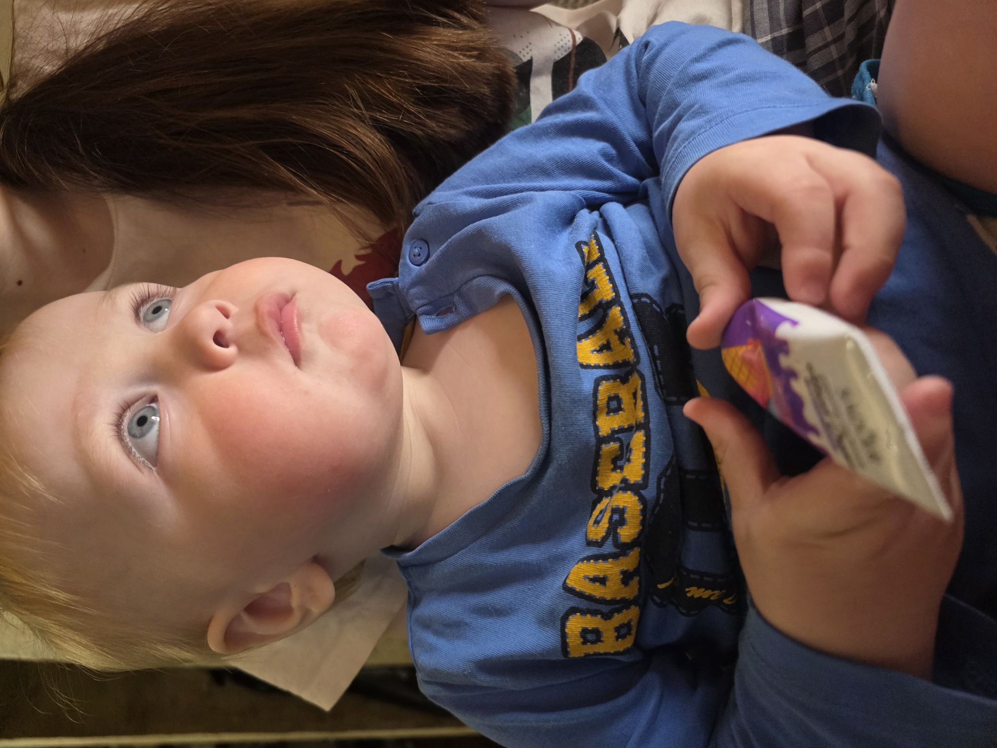 A baby with blond hair and blue eyes is lying on their back. The baby is wearing a blue long-sleeved shirt with yellow and black lettering. The baby's right hand holds a white tube with purple and yellow designs. Long brown hair is visible above the baby's head. The background consists of various fabrics and materials, including what appears to be a white cloth or sheet. It is not possible to determine the location.