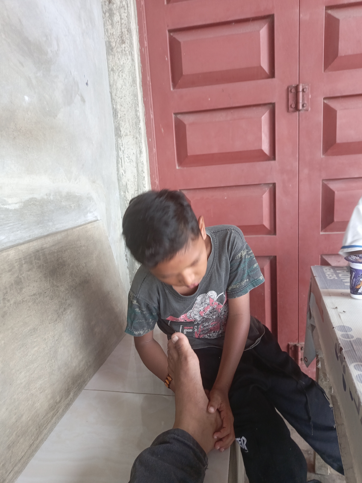 The image depicts a young boy sitting indoors, looking down at his foot which is held by another person. The boy is wearing a dark t-shirt with a graphic print and dark pants. His foot has an orange bracelet on the ankle. The background shows a red, paneled door and a light-colored wall. To the right, a table is visible with a cup on it and some ants on the surface. The floor is tiled. The scene appears to be in a residential or public space in Lhokseumawe, Indonesia. The lighting suggests it is daytime.