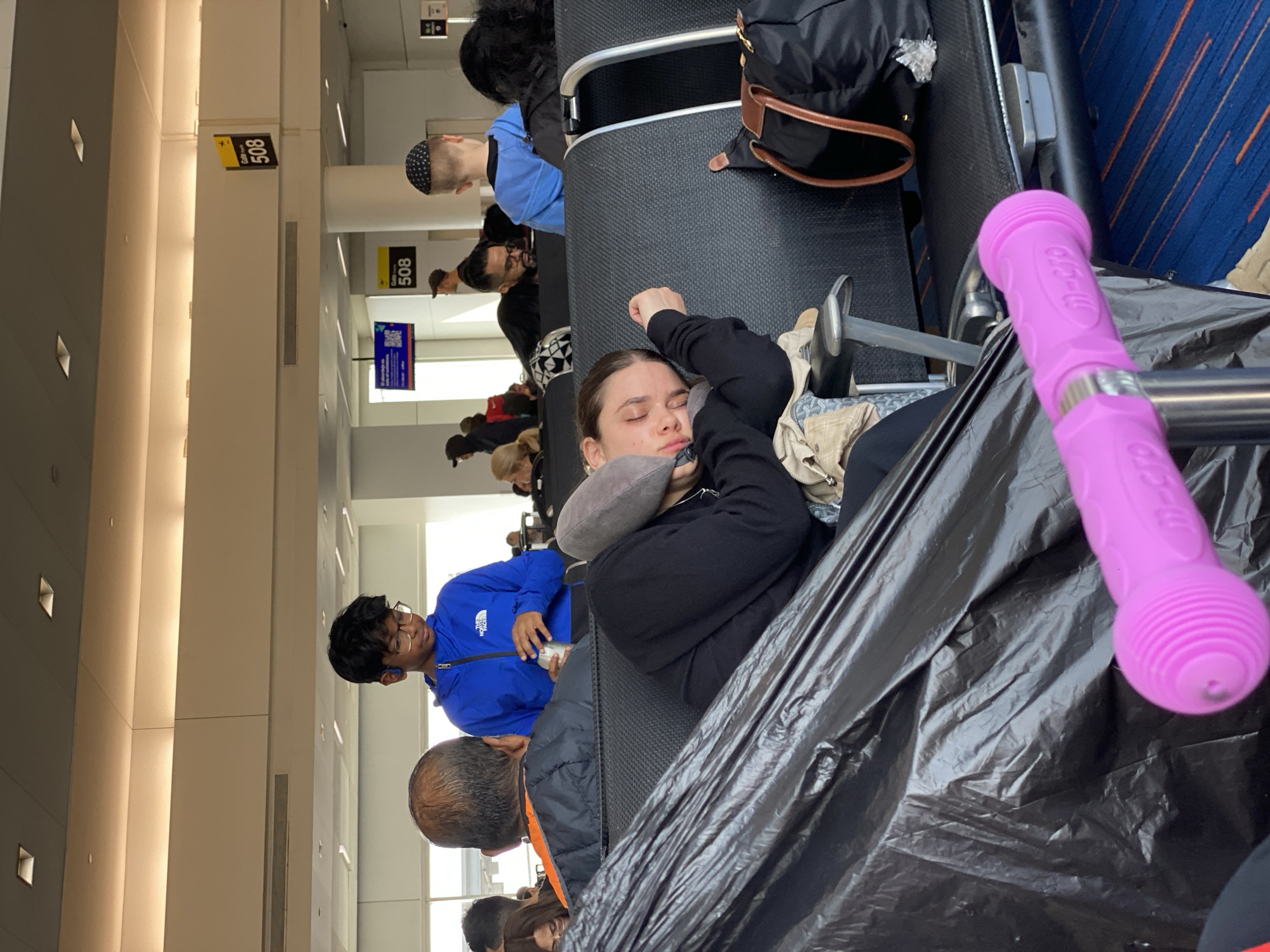 The image depicts an airport waiting area in New York City, United States of America, where people are waiting for their flights. A woman sleeps on a seat with a neck pillow. Other passengers are seated, some looking at their phones or talking. A sign indicates gate 508. A pink child's scooter is present in the foreground. The overall environment suggests a busy airport atmosphere.