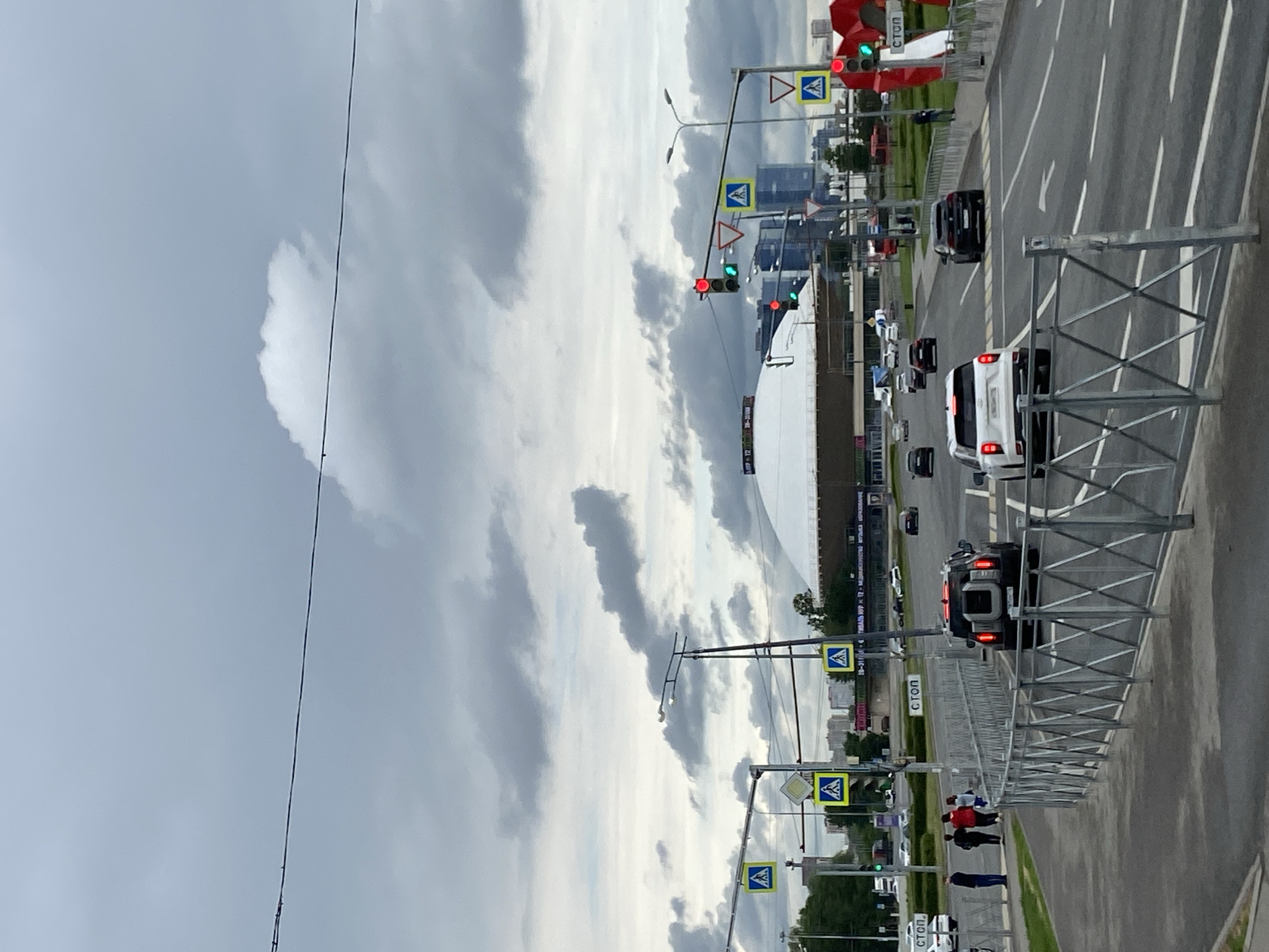 The image shows a view of Kazan, Russia, featuring a road with traffic and pedestrian walkways. There are several cars driving on the road, including a white SUV and a black vehicle. Pedestrians are present on the side of the road near pedestrian crossings, which are marked with blue and yellow signs. Traffic lights can be seen along the road, some displaying red and green signals. The Kazan Arena stadium is visible in the background, indicating the location. The sky is overcast with various cloud formations. Metal guardrails line the side of the road.
