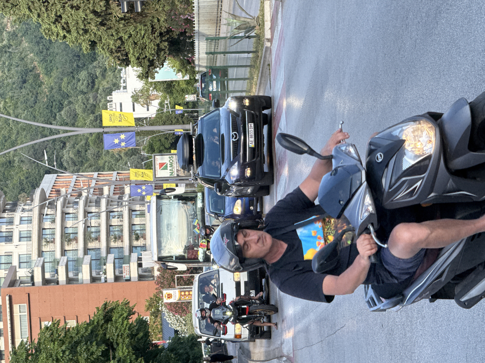 In Budva, Montenegro, a man on a scooter rides in traffic on a sunny day. The man, wearing a black t-shirt and shorts, has a helmet on his head. He is riding a dark grey scooter. In the background, cars, a white van, and a bus are visible. Buildings and trees line the street. The image appears to be taken near a pedestrian crossing.