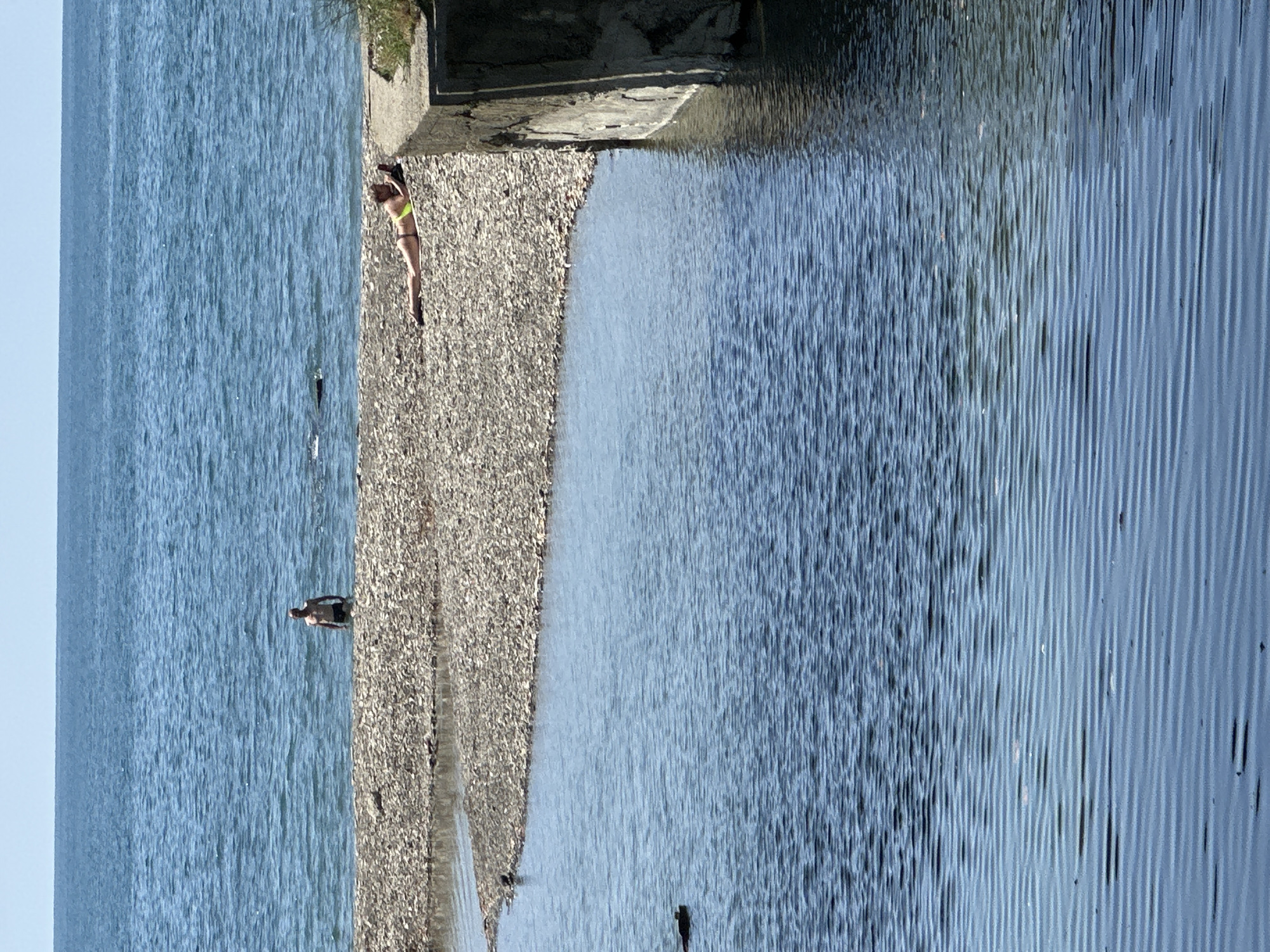 An overhead shot captures a scene of leisure at a beach in Sochi, Russia. The foreground is dominated by rippling blue water, possibly a river or a sheltered cove, with a dark object barely visible on the surface towards the left. Beyond this, a pebble beach separates the water from a vast expanse of blue sea under a clear sky. On the pebble beach, a man stands partially submerged in the sea, facing away from the camera. Further to the right, a woman lies on her stomach on the beach, wearing a yellow bikini and looking at her phone. A concrete structure runs along the right edge of the beach.