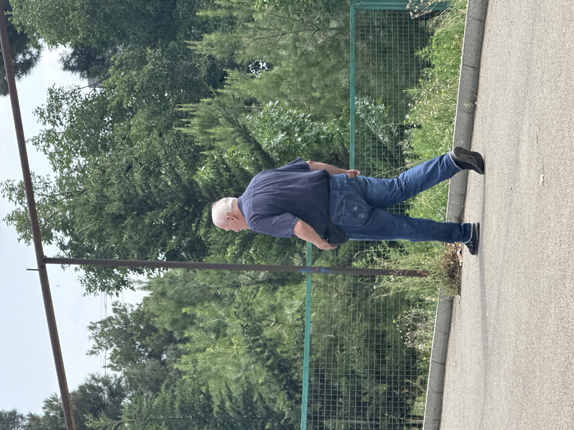 A middle-aged Caucasian male is walking away from the camera on a paved path in a park-like setting. He is wearing a navy blue short-sleeved collared shirt, blue jeans, and dark sneakers. He has a dark fanny pack or belt bag around his waist. The background consists of lush green trees and a metal fence. The sky is overcast. The location is identified as Tbilisi, Georgia.