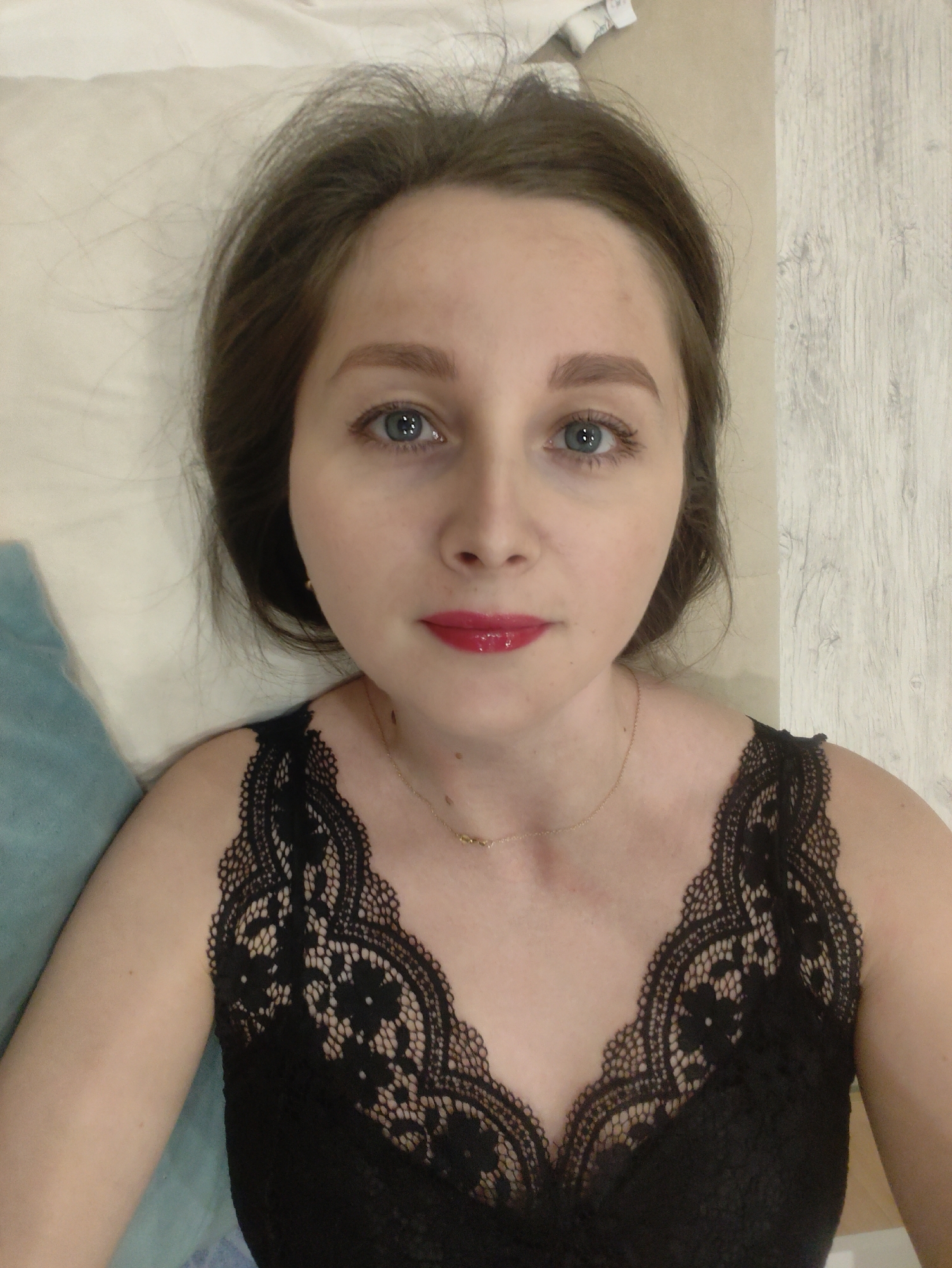 A young woman with light skin and brown hair is lying down, facing the camera. She has blue eyes, light eyebrows, and is wearing red lipstick. She is wearing a black lace top with a V-neck. A thin gold necklace is visible around her neck. She is lying on a white surface, possibly a bed, with a blue pillow visible on the left side of the frame. The background behind her head appears to be a light-colored wall or possibly a wooden floor. The location is unknown.
