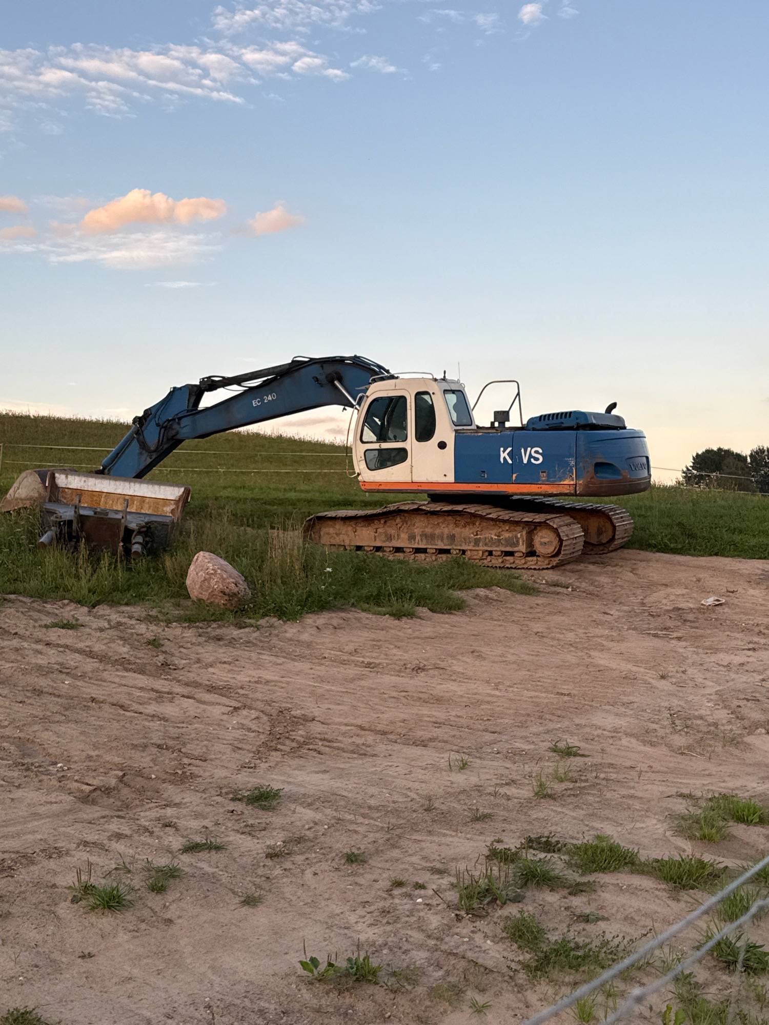 In the city of Galinė, Lithuania, an excavator with the markings "EC 240" and "KVS" is positioned on a dirt and grass terrain. The excavator is blue and white with orange trim. A trailer with a rusty bucket attachment is to the left of the excavator, partially obscured by tall grass. A large, smooth boulder rests on the ground between the trailer and the excavator. The background consists of a grassy incline beneath a pale blue sky with scattered, pink-tinged clouds. A thin wire fence is visible in the lower right corner of the image. The scene appears to be outdoors, possibly at a construction site or a storage area for heavy machinery.