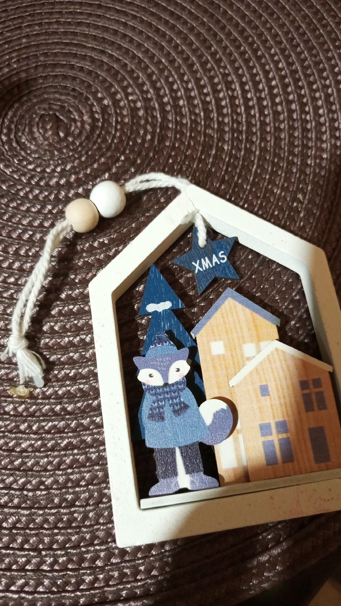 The image shows a Christmas tree ornament on a textured brown surface. The ornament is a white frame in the shape of a house, containing a blue fox wearing a hat and scarf, two blue Christmas trees, a tan house, and a blue star labeled "XMAS". The ornament hangs from a white string with two wooden beads. There is no indication of this image being taken in the city of Юдино, Russia.