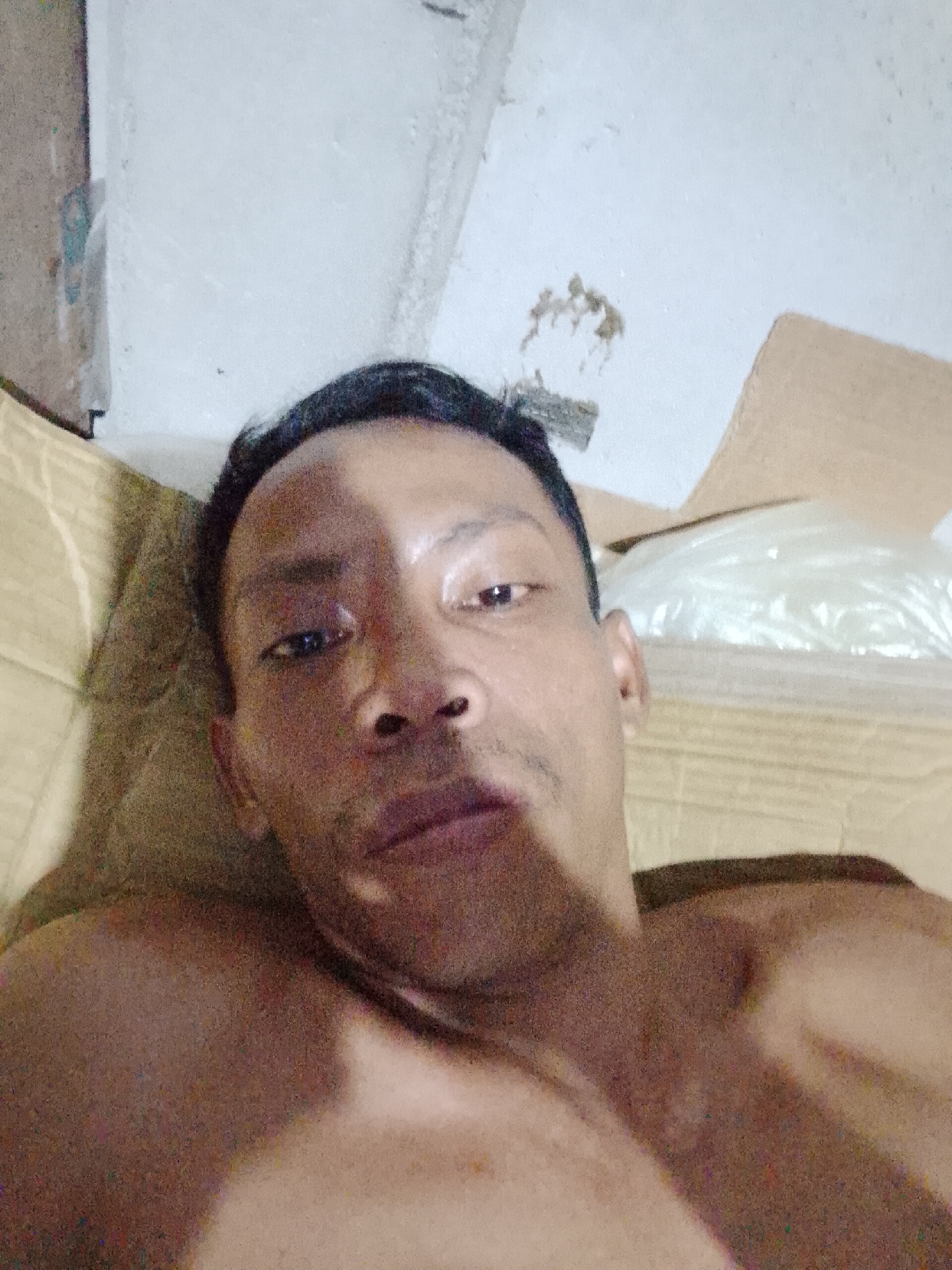 A close-up, low-angle photograph depicts a shirtless man with short, dark hair lying on a tan, cushioned surface. The man is facing the camera, his head slightly tilted. His complexion is brown, and he has a mustache and a dark shadow on the left side of his face. Behind him are cardboard boxes and a translucent plastic bag. The background also includes a white wall with minor damage and discoloration. The photograph was presumably taken indoors, but the location within Bukit Ketok, Indonesia, cannot be definitively ascertained from the image alone.