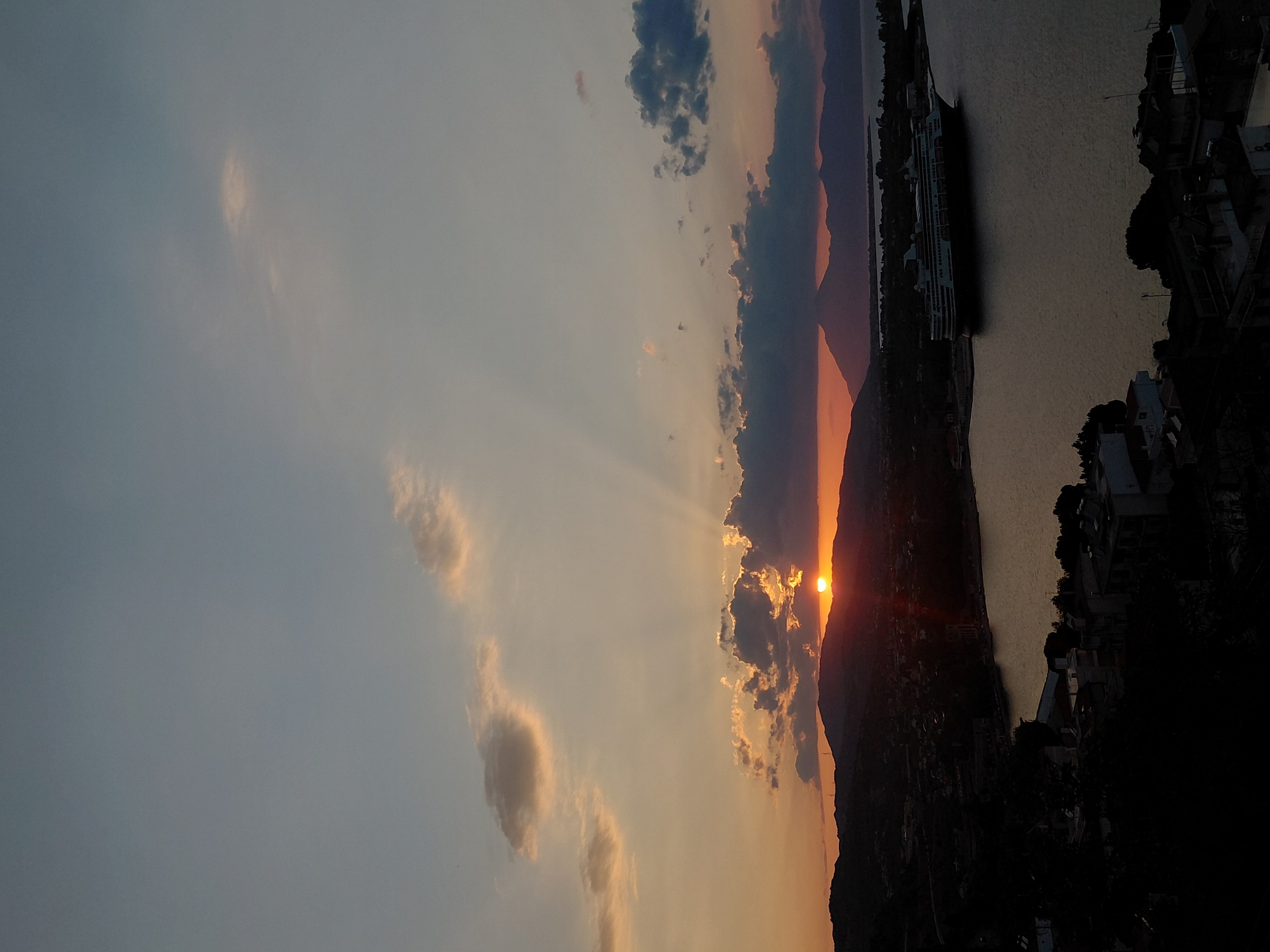The image depicts a sunset over a coastal area in the Municipal Unit of Aegio, Greece. The sun is partially obscured by clouds, casting rays of light across the sky. A large cruise ship is docked in the harbor, with a town and hills visible in the background. The foreground is dominated by silhouetted buildings, suggesting an elevated viewpoint. The overall scene conveys a peaceful and picturesque atmosphere.