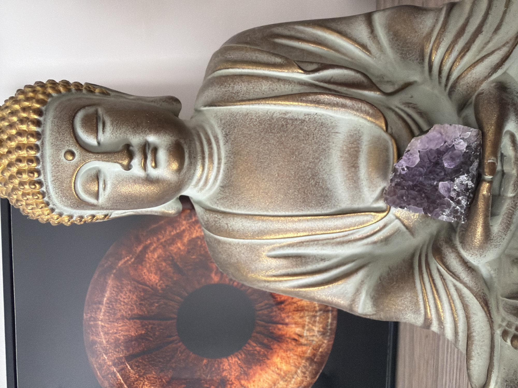 This media file features a close-up, horizontal shot of a Buddha statue. The statue is predominantly light beige with metallic gold accents, particularly on the hair, facial features, and the folds of its robes. It appears to be made of resin or a similar material, with a slightly textured finish. Resting on the statue's chest, near its hand, is a cluster of purple amethyst crystals, adding a natural, vibrant element to the otherwise subdued tones of the statue.
The background consists of a plain white surface on the top right, suggesting an indoor setting like a home or studio. To the lower left and partially obscured by the statue, there is a framed image or artwork. This artwork features a close-up of an iris, rendered in warm, earthy tones of brown and orange, with a dark pupil at its center. The overall composition creates a serene and contemplative atmosphere. There are no discernible cues for time of day or weather. No visible text is present in the image. The setting can be inferred to be within Nha Trang Ward, Vietnam, based on the provided location context. The activity, if any, is static, focusing on the presentation of the Buddha statue and the amethyst.