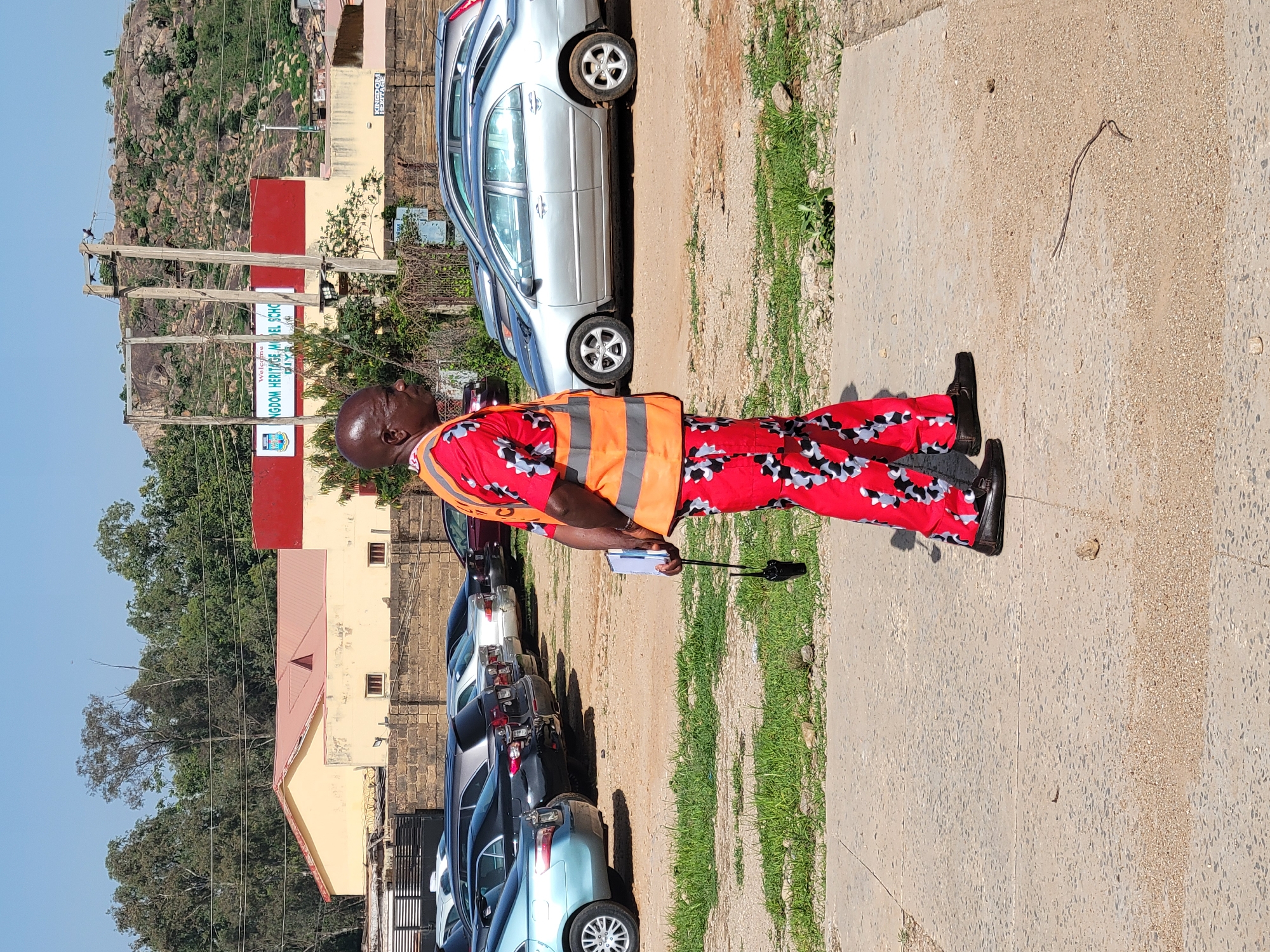 A man stands on a concrete surface, wearing a red and black patterned shirt and pants, black shoes, and an orange traffic vest. He is holding a black, t-shaped object and a clipboard. Behind him is a row of parked cars and buildings. A sign indicates "KINGDOM HERITAGE MODEL SCHOOL." A hill or mountain is visible in the background. The setting is Gura Top, Nigeria.