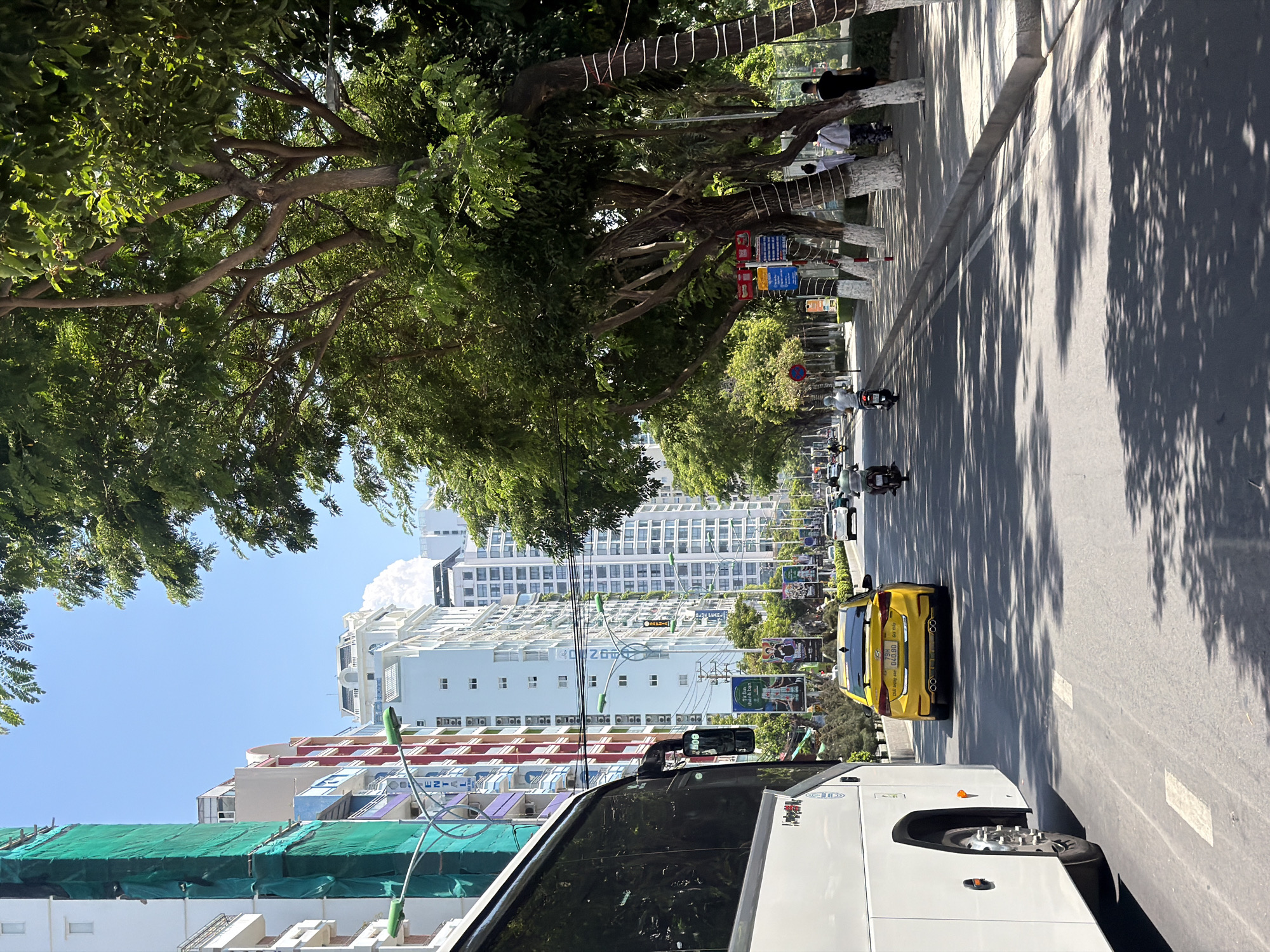The image depicts a sun-drenched street scene in Nha Trang Ward, Vietnam, with high-rise buildings lining the right side of the road and lush trees providing shade along the sidewalk. A large white tour bus is prominently featured on the left side, partially obscuring the view of the buildings which display a mix of architectural styles and vibrant colors, some undergoing construction with green netting. A bright yellow taxi is visible in the middle ground, driving on the right side of the road, preceded by several motorbikes and followed by other vehicles. Pedestrians are seen walking on the sidewalk, some under the shade of the trees. The road is paved, with clear lane markings, and the sky is a bright blue with a few wispy clouds. The overall impression is one of a bustling urban environment on a clear day.