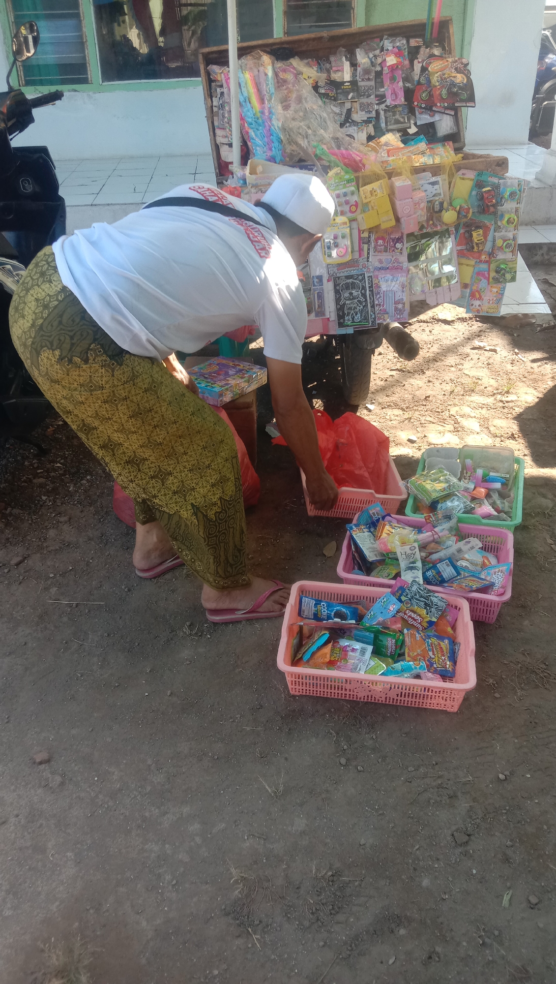 A person, wearing a white t-shirt, a white peci hat, and a patterned sarong, is bent over a small mobile cart loaded with various toys and snacks. The cart is positioned on a dirt ground next to a paved area. In front of the cart, there are at least four plastic baskets filled with individually packaged snacks. The person appears to be arranging or selecting items from the cart. The background shows a building with windows and a motorcycle parked to the left. The scene is likely outdoors in a public area, possibly a street market or roadside stall. The location is identified as Ngempit, Indonesia.