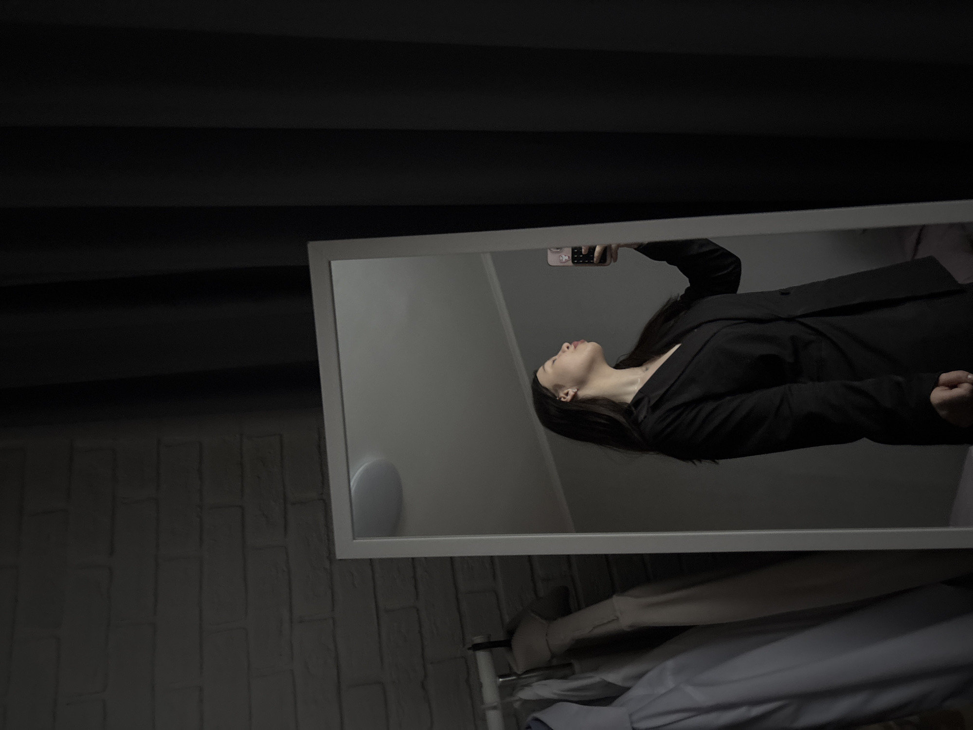 A medium shot captures a reflective scene within a white-framed mirror. A woman, wearing a black blazer, is holding a phone up to take a photo of herself, her face visible in profile. The mirror's reflection shows an interior space with a pale grey wall and a round speaker. Outside the mirror's reflection, the environment consists of a dark curtain above, a textured brick wall to the left, and clothes hanging on a rack below the mirror. The location is Perm, Russia.