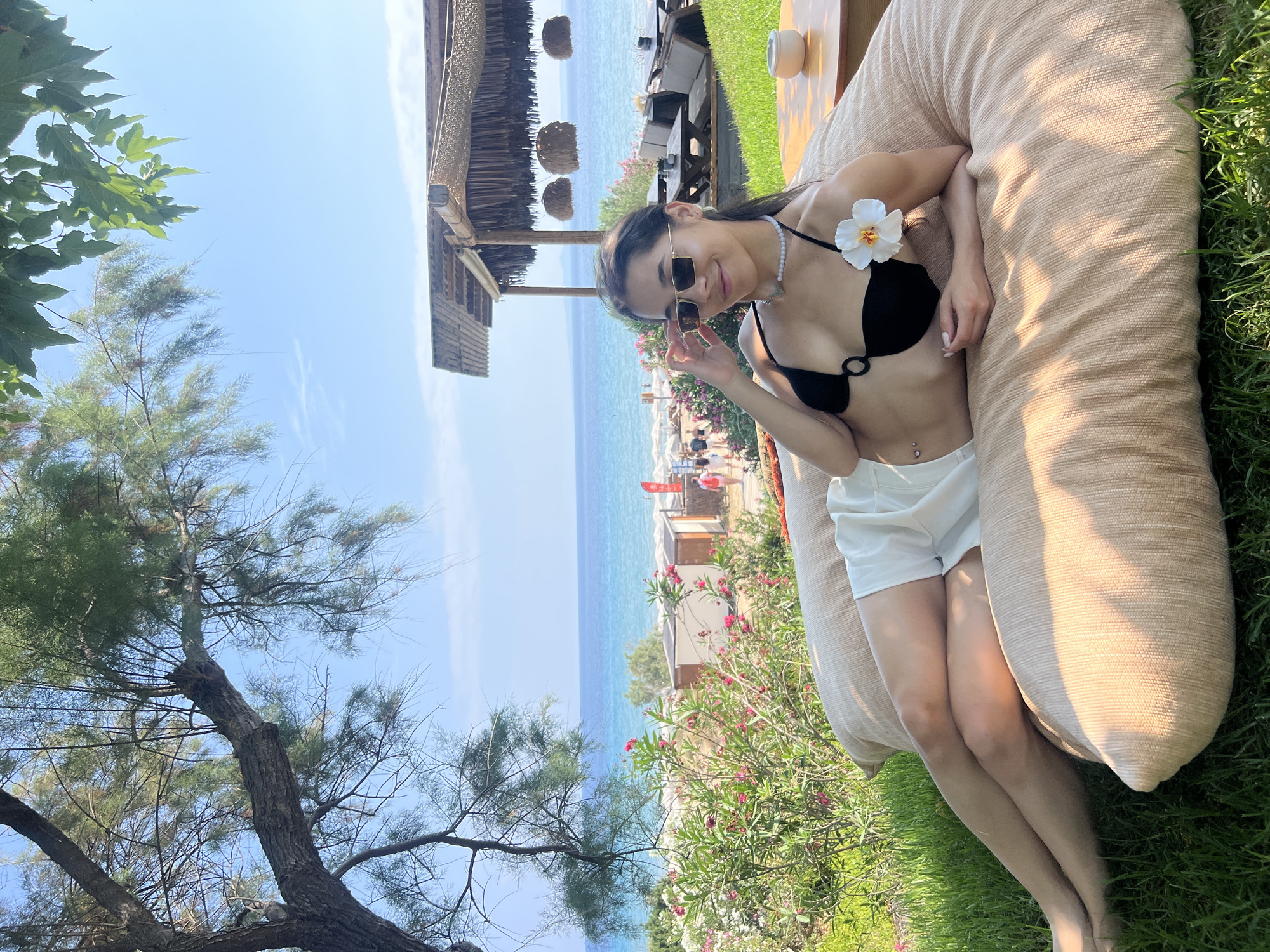A young woman with long dark hair, wearing a black bikini top, white shorts, sunglasses, and a pearl necklace, reclines on a large tan floor cushion outdoors on a grassy area. She has a white flower pinned to her bikini strap. In the background, a beach with turquoise water and blue sky is visible. A thatched-roof structure with tables and chairs is located to the right of the woman. The setting appears to be a beachside resort or restaurant in Zakynthos Municipal Unit, Greece, likely during daylight hours given the bright sunlight and clear sky.