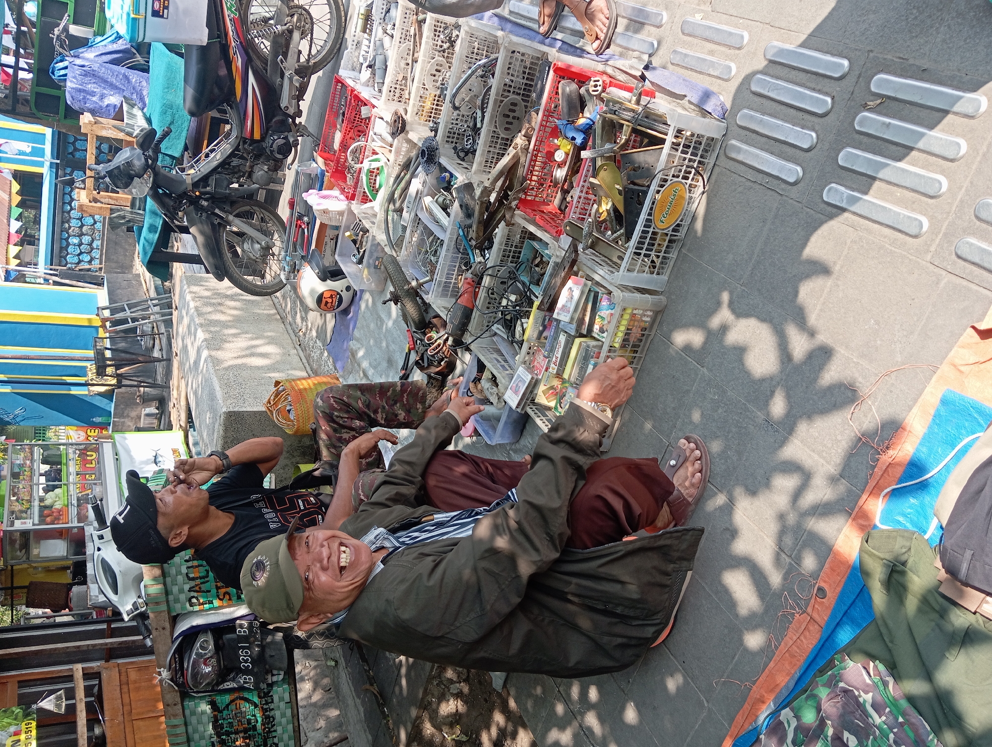 A street vendor in Borobudur, Indonesia, sits crouched beside a collection of items laid out on the ground, including various tools and motorcycle parts displayed in plastic crates. He is wearing a green cap and a dark jacket over a striped shirt and dark pants. To his right, another man sits, wearing a black cap and t-shirt, smoking. A motorcycle is parked behind them. The scene appears to be set up on a sidewalk next to a street, with some stalls or shops visible in the background.