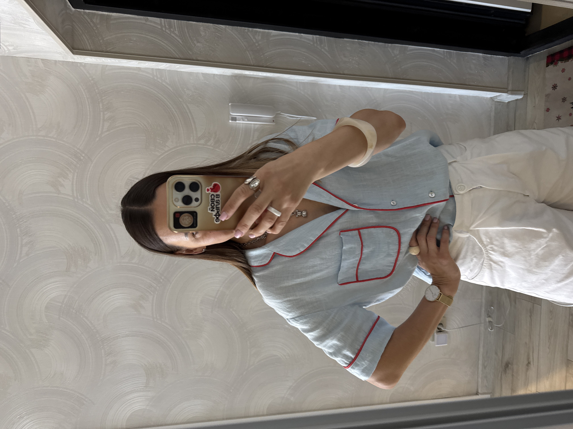 A person is taking a mirror selfie in a room. The person is wearing a light blue short-sleeved pajama-style shirt with red trim and white wide-leg pants. They are also wearing a watch on their left wrist and rings on their right hand. The room has patterned wallpaper and a wooden floor. The image does not provide any information about the city of Sochi, Russia.