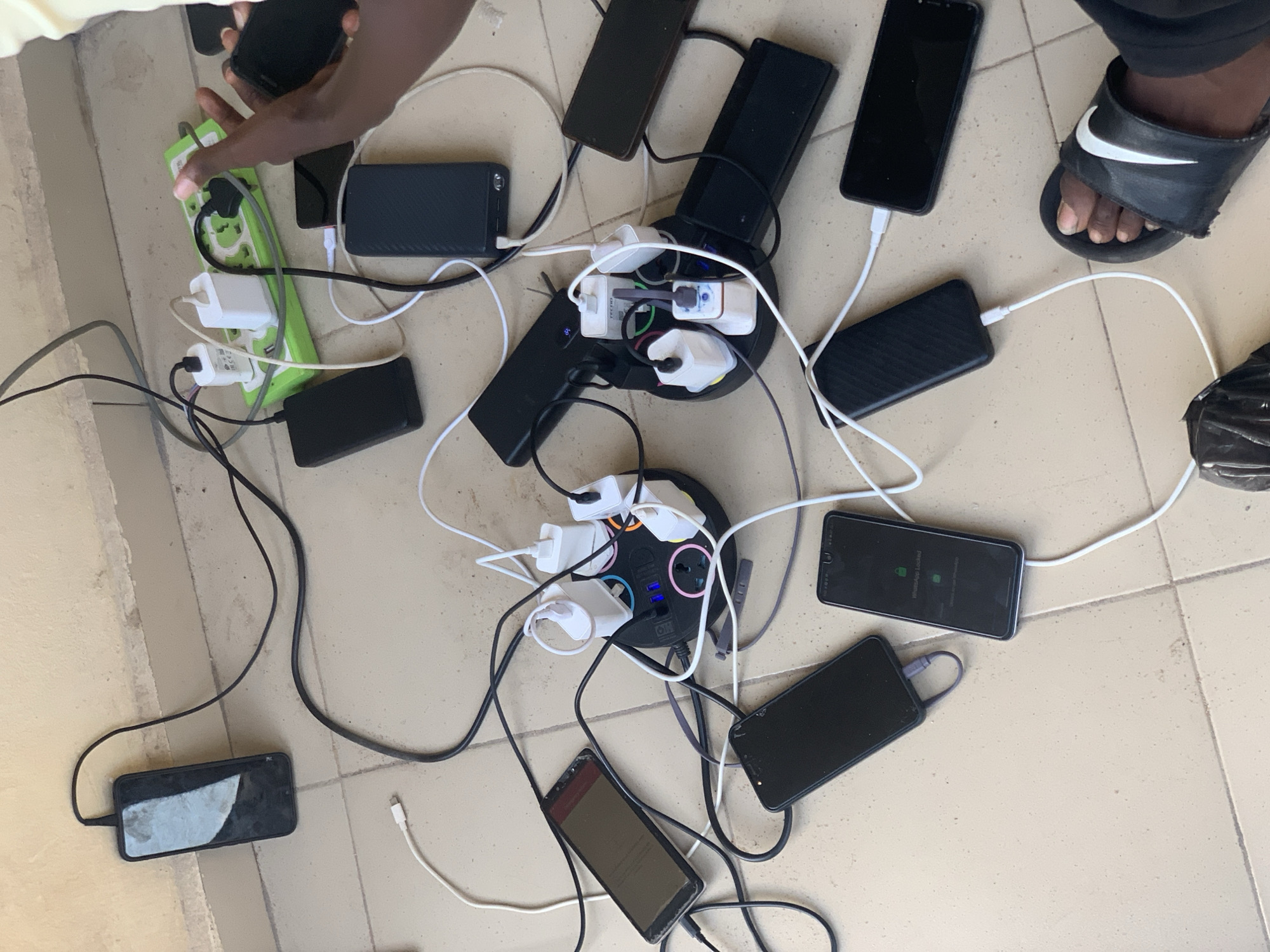 The image captures a busy scene of multiple smartphones being charged simultaneously on a light-colored tiled floor, likely indoors or in a covered area, in Lokoja, Nigeria. At least ten smartphones are scattered across the floor, all connected to various power sources.
The central activity involves extensive device charging. One person's hands are visible at the top left, actively plugging a black smartphone into a bright green power strip that has multiple outlets and white charging bricks. Another person's feet, wearing a black Nike slide sandal with a white swoosh logo, are visible on the right side of the frame.
Several power strips and extension cords are in use. A prominent black circular power strip with blue indicator lights is visible in the center, accommodating multiple white charging bricks and USB cables. Another long, green power strip is seen on the left. Numerous black and white USB cables crisscross the floor, connecting phones to the power sources. Several power banks are also visible, some acting as charging hubs themselves or being charged.
Notable details include text visible on some devices: one phone screen clearly displays "WhatsApp Locked" with instructions to "Touch the fingerprint sensor or enter pattern to unlock." Another phone screen shows what appears to be a web page or application, though the text is not entirely legible. The CE mark is visible on at least one white charging brick. The overall impression is one of a communal or shared charging station, possibly due to limited access to power outlets or a need to charge many devices at once.