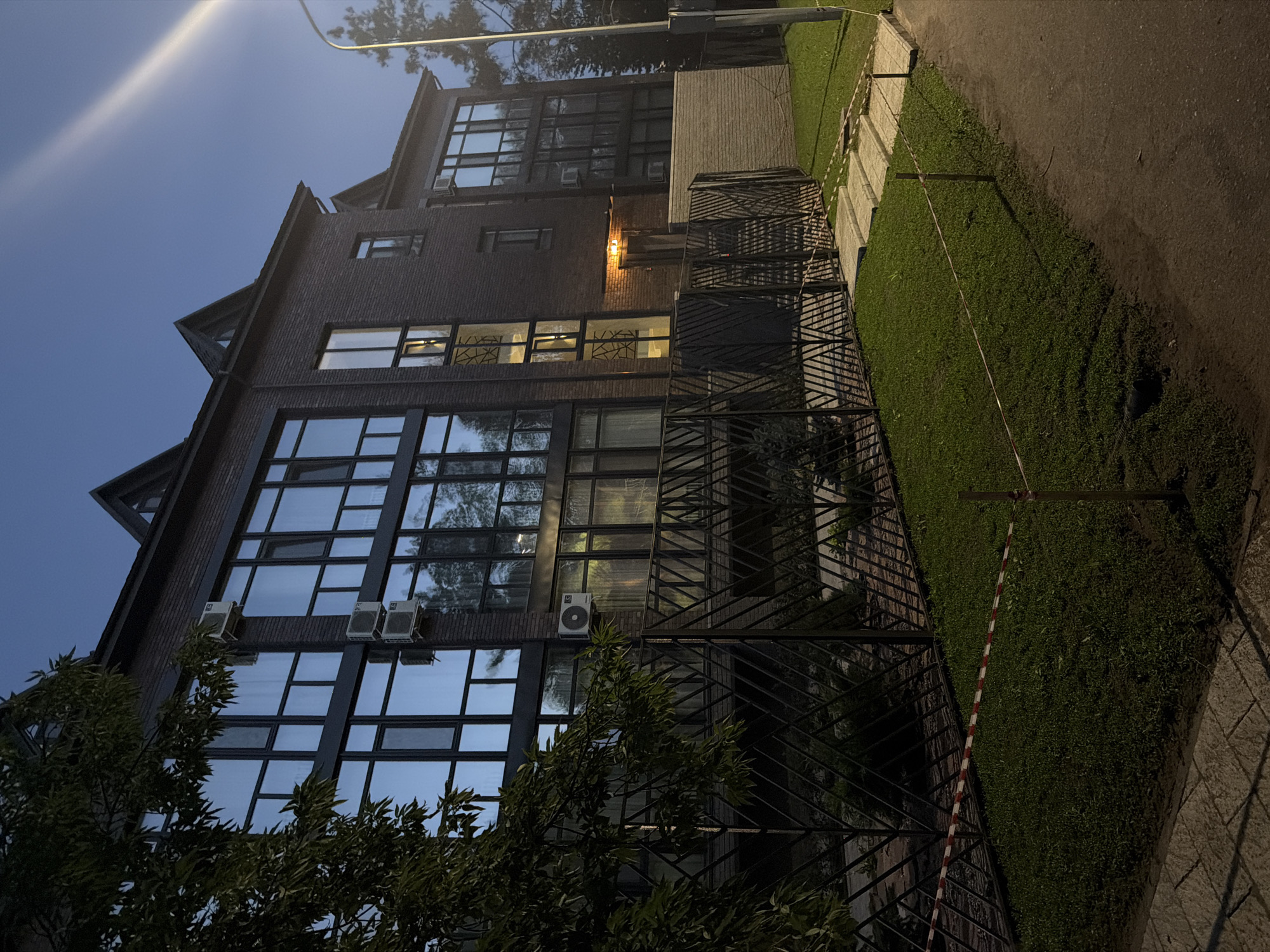 The image shows a modern, multi-story brick building with large, dark-framed windows. The building appears to be residential or office space. Several outdoor air conditioning units are visible mounted on the exterior walls. A black metal fence with a geometric pattern runs along the front of the property, separating it from a grassy area. There are some steps leading up to an entranceway, which is illuminated by an overhead light. Red and white caution tape is strung across the lawn in front of the fence, supported by wooden posts. The scene is set at dusk or at night, with artificial lighting visible both from within the building and from a streetlamp on the right side of the image. The sky is a deep blue, suggesting either twilight or nighttime. The surrounding environment includes trees and a paved walkway or road. The location is identified as Pavlodar, Kazakhstan.