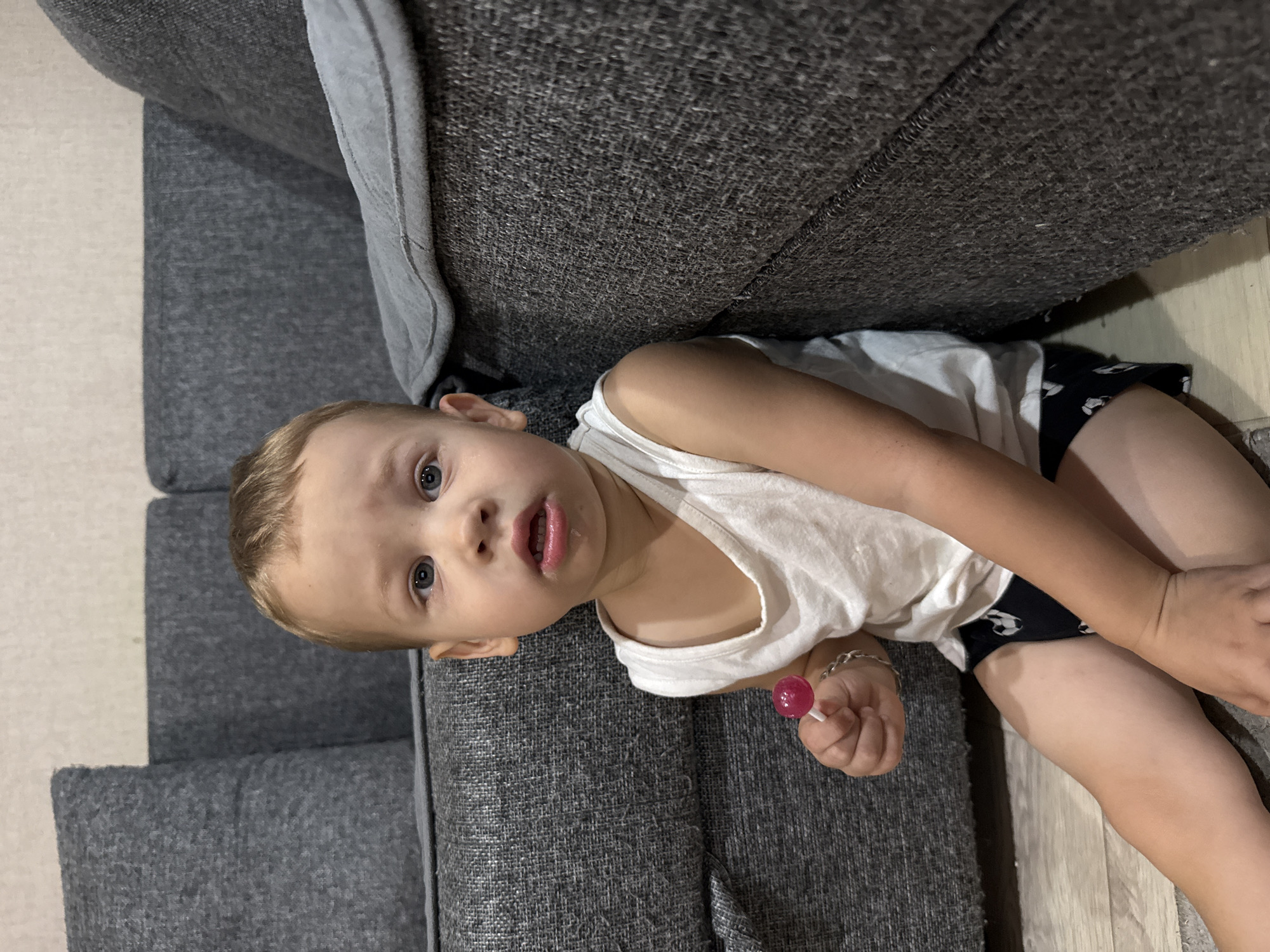 A close-up, waist-up shot shows a young boy lying on a grey couch. He is wearing a white tank top and black shorts with a white pattern. His right arm is extended, and he is holding a small, pink lollipop. The boy has blonde hair and blue eyes, and he is looking directly at the camera with a slightly open mouth. His expression appears curious or inquisitive. The couch fabric is textured, and a sliver of light-colored flooring is visible to the right of the boy. The lighting suggests it is daytime, possibly indoors. The overall mood is casual and candid. There is no visible text in the image.