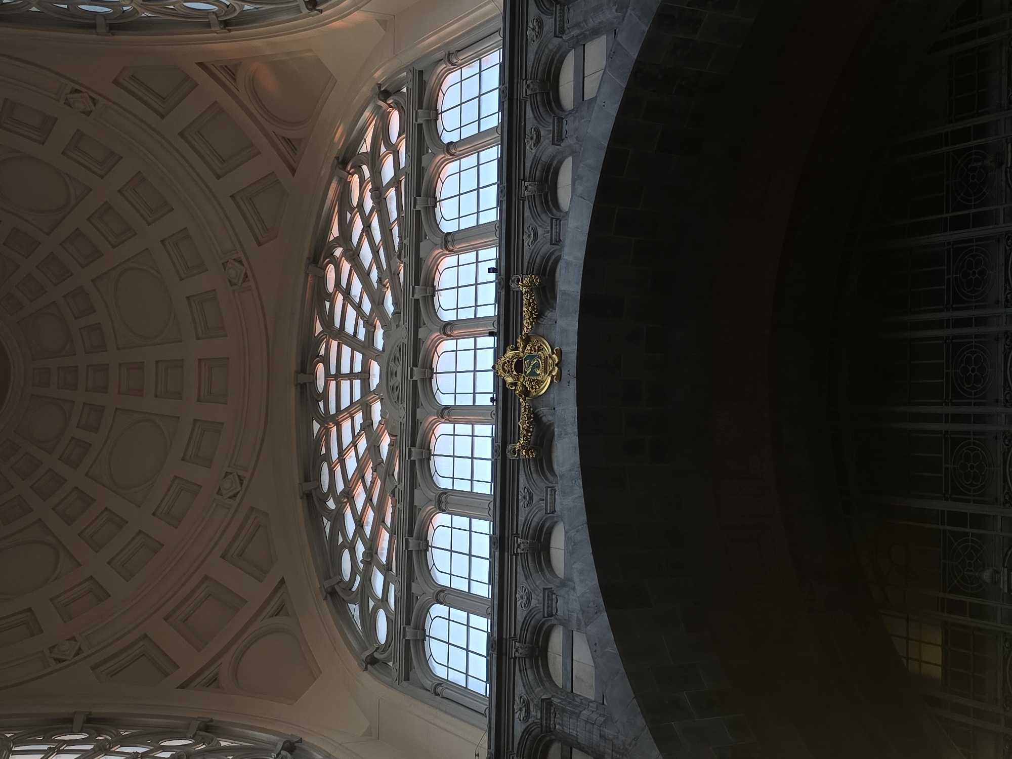 The image captures a low-angle interior view of a grand architectural space, likely a train station, in Antwerp, Belgium. The dominant features are a large, ornate arched window with intricate tracery, set above a series of smaller arched windows. Above these, the ceiling is a coffered dome with geometric patterns. Below the arched windows, a dark, massive stone archway frames a view of decorative metalwork and the ceiling of a lower level. A golden crest adorns the stone facade between the arched windows. The lighting suggests either natural light filtering through the windows, casting warm tones on the ceiling, or artificial lighting highlighting the architectural details. The scene is devoid of people, focusing solely on the impressive construction and design of the building. The architecture suggests a historical period, possibly late 19th or early 20th century, characterized by neoclassical or eclecticism influences.