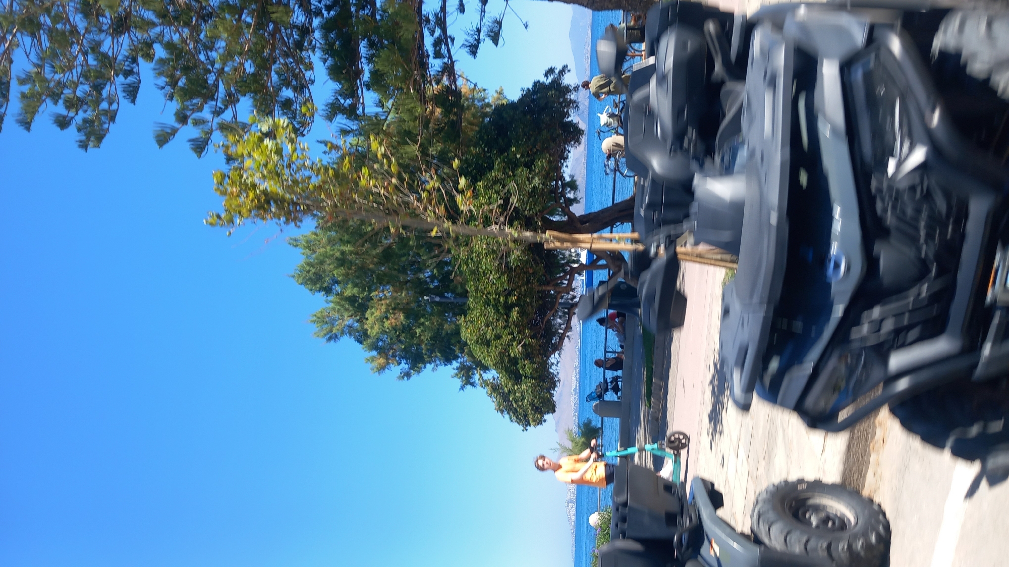 A young man in an orange t-shirt rides a teal electric scooter along a paved walkway next to a body of water. In the foreground, several dark-colored ATVs are parked or being transported. Behind the scooter, a line of people are seated on a low wall facing the water. A large tree with lush green foliage stands behind them, and in the distance, islands and a clear blue sky are visible. The scene appears to be in Kos, Greece, likely along the waterfront promenade of Kos town, a popular area for tourists and locals.