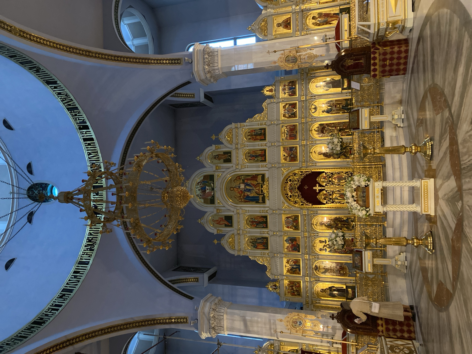 The image depicts the interior of a church, likely located in Saint Petersburg, Russia, judging by the style of the architecture and religious iconography. The interior is ornate, with a large, gilded iconostasis filling the frame. This iconostasis is segmented into multiple tiers, each containing religious paintings or icons. A large, gold-colored chandelier hangs from the ceiling, partially obscuring the view. The ceiling is vaulted and decorated with inscriptions. Marble columns and pilasters flank the iconostasis. In the foreground, a person is seen standing in front of the iconostasis.