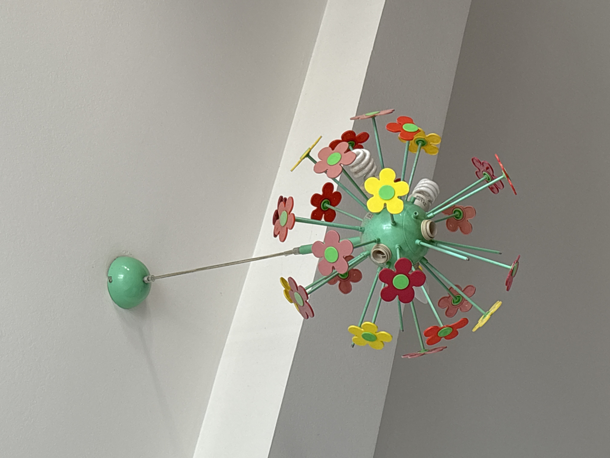 The image shows a colorful, flower-shaped light fixture hanging from a white wall. The fixture has a light green base with multiple green rods extending outwards, each topped with a flower in varying colors (red, pink, yellow, and orange) with green centers. Some rods have screw-in light bulbs. The fixture is mounted to the wall via a light green sphere attached to a thin metal rod. The wall is a neutral white color and forms an arch in the background, suggesting an interior space. The exact location in Kragujevac, Serbia, cannot be determined from the image alone.