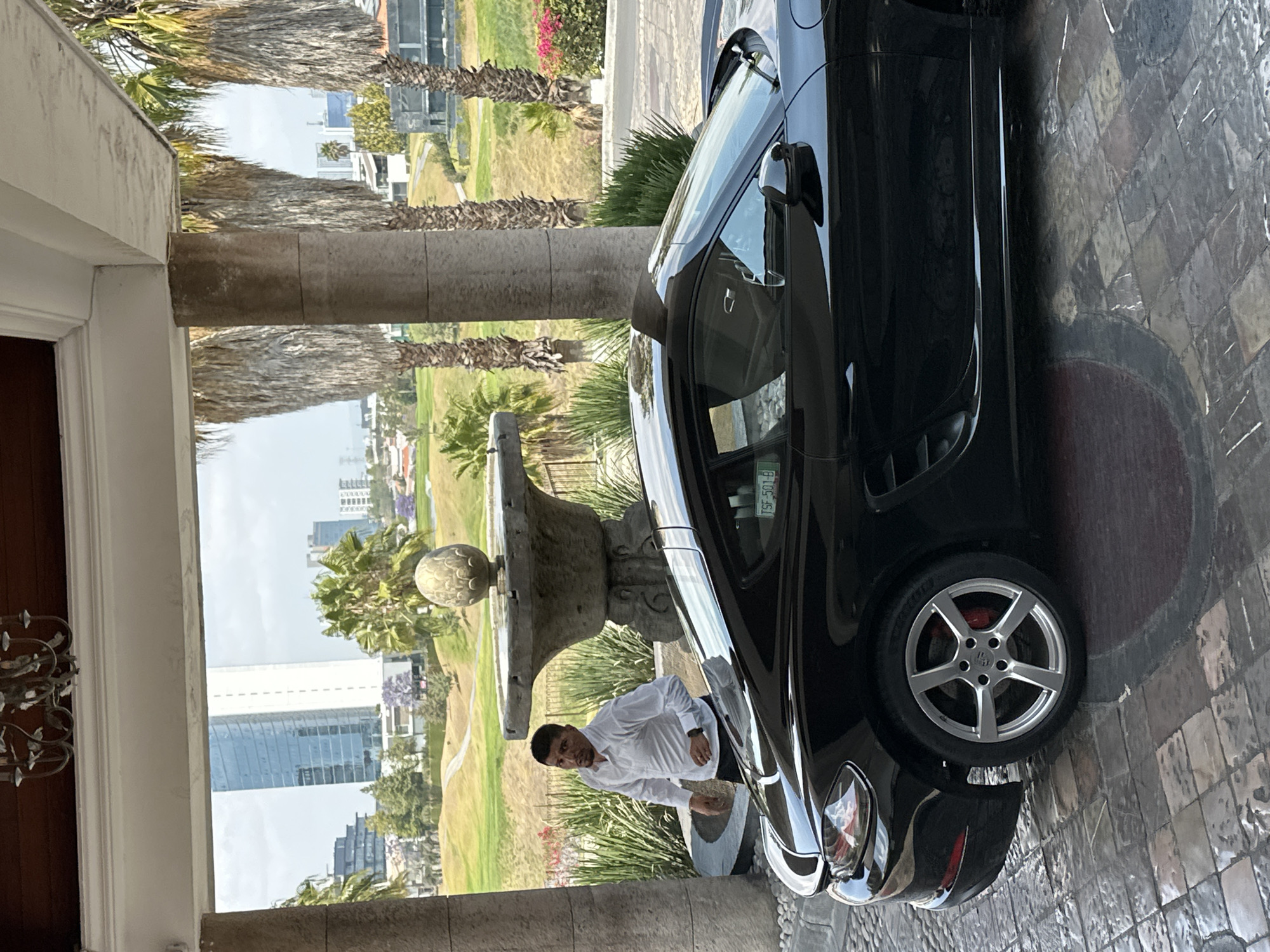 A man in a white shirt stands near a black Porsche in front of a stone fountain. The fountain and Porsche are positioned near the entrance of a building supported by stone pillars. Beyond the pillars is a view of a golf course with palm trees and buildings in the background, possibly in San Bernardino Tlaxcalancingo, Mexico. The ground near the car is paved with stone tiles.
