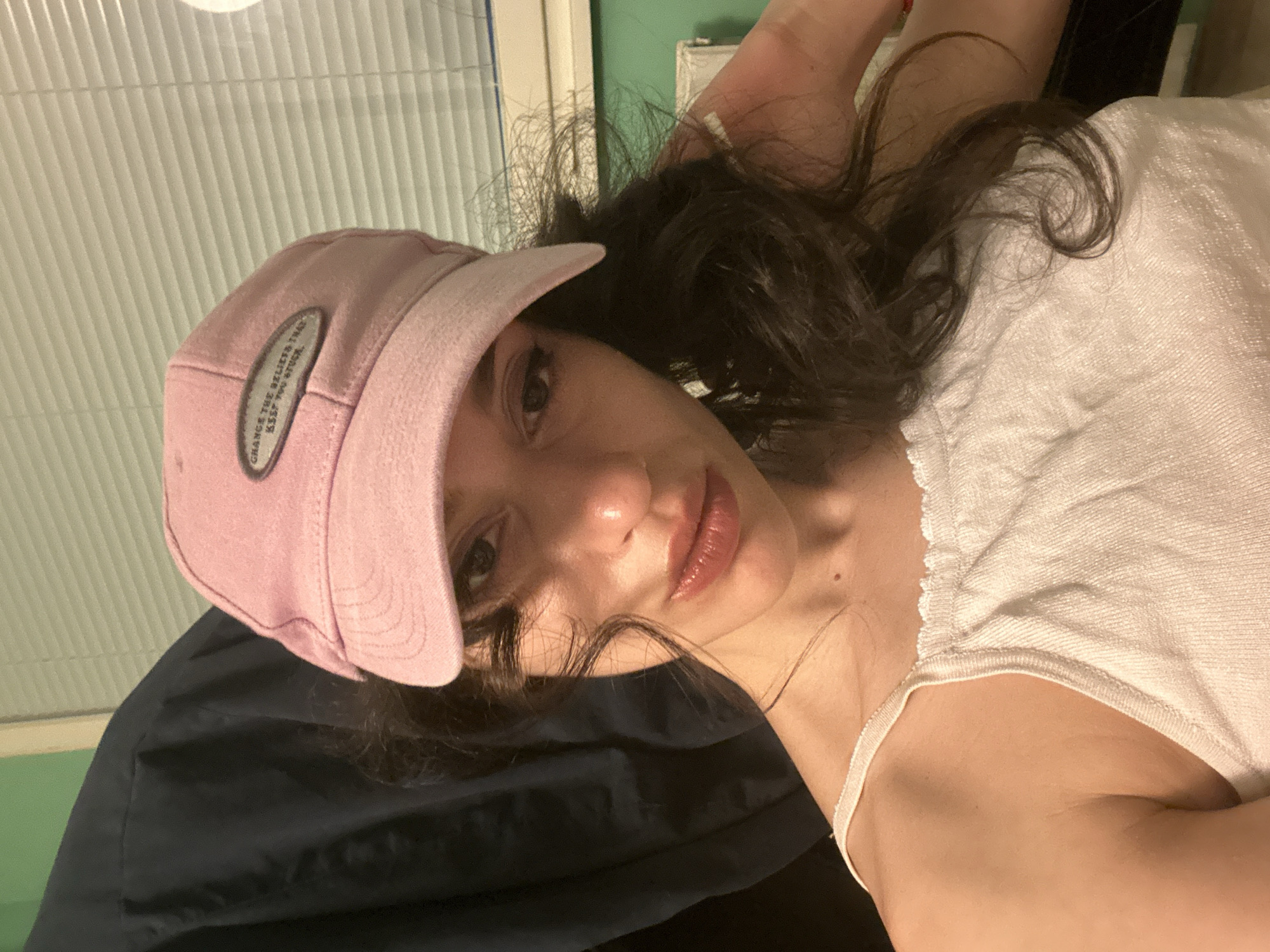 A young woman with dark, curly hair and light skin is taking a selfie. She is wearing a pink baseball cap and a white camisole. The cap has a patch with text on it. Her makeup includes lipstick and eyeliner. The background is an indoor setting with blinds on a window and a dark garment behind her. There is a faint light source to her right. The image is a close-up, focusing on her face and upper body. No specific location in Sofia, Bulgaria is indicated in the image.