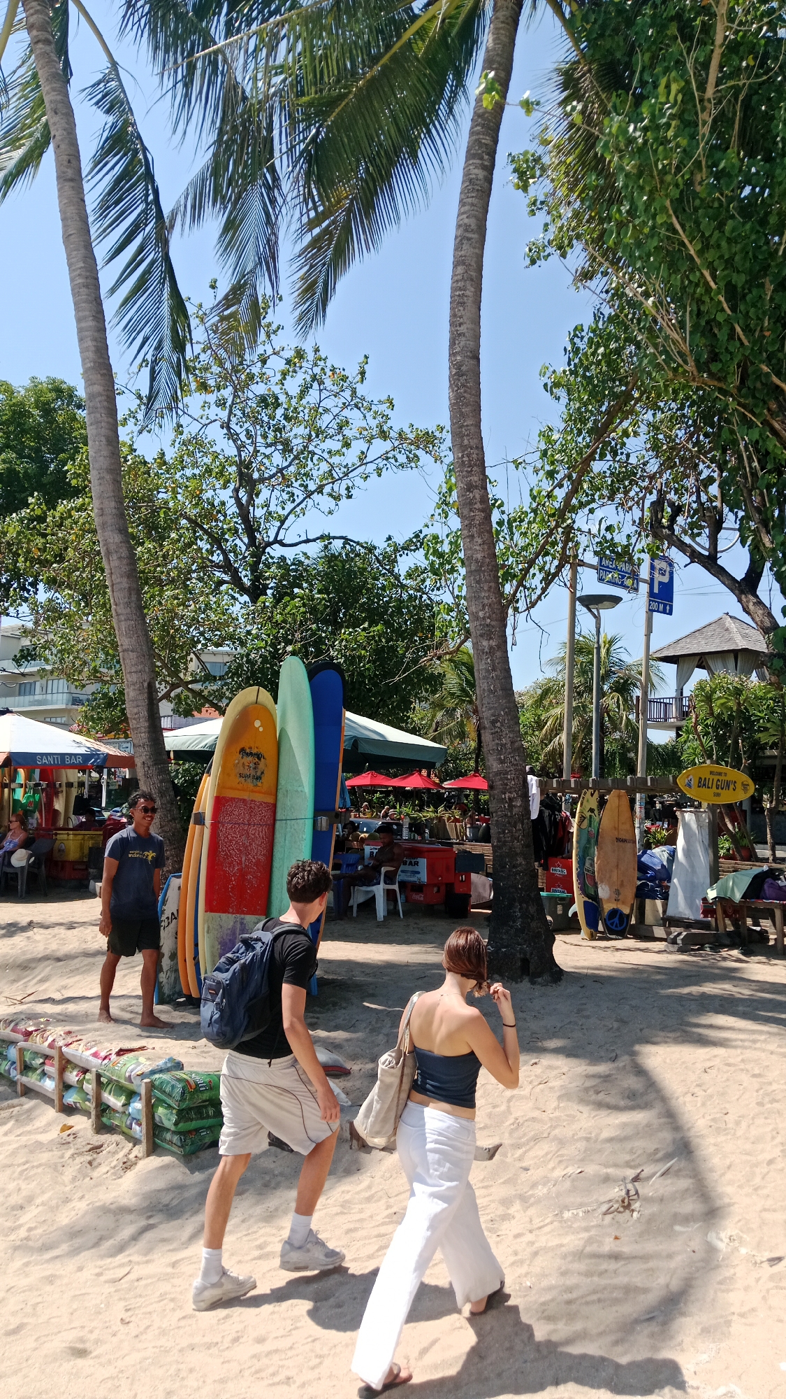 The image depicts a beach scene with several palm trees. In the foreground, two people are walking on the sand; a man in a black t-shirt and white shorts with a blue backpack, and a woman in a blue tube top and white pants. Behind them, a collection of surfboards are propped upright. Further back, there are market stalls with umbrellas and tables, and more surfboards. In the background, a building with a balcony is visible, and trees line the area. A sign that says "WELCOME TO BALI GUN'S" is visible on the right side of the image. The sky is clear and blue.