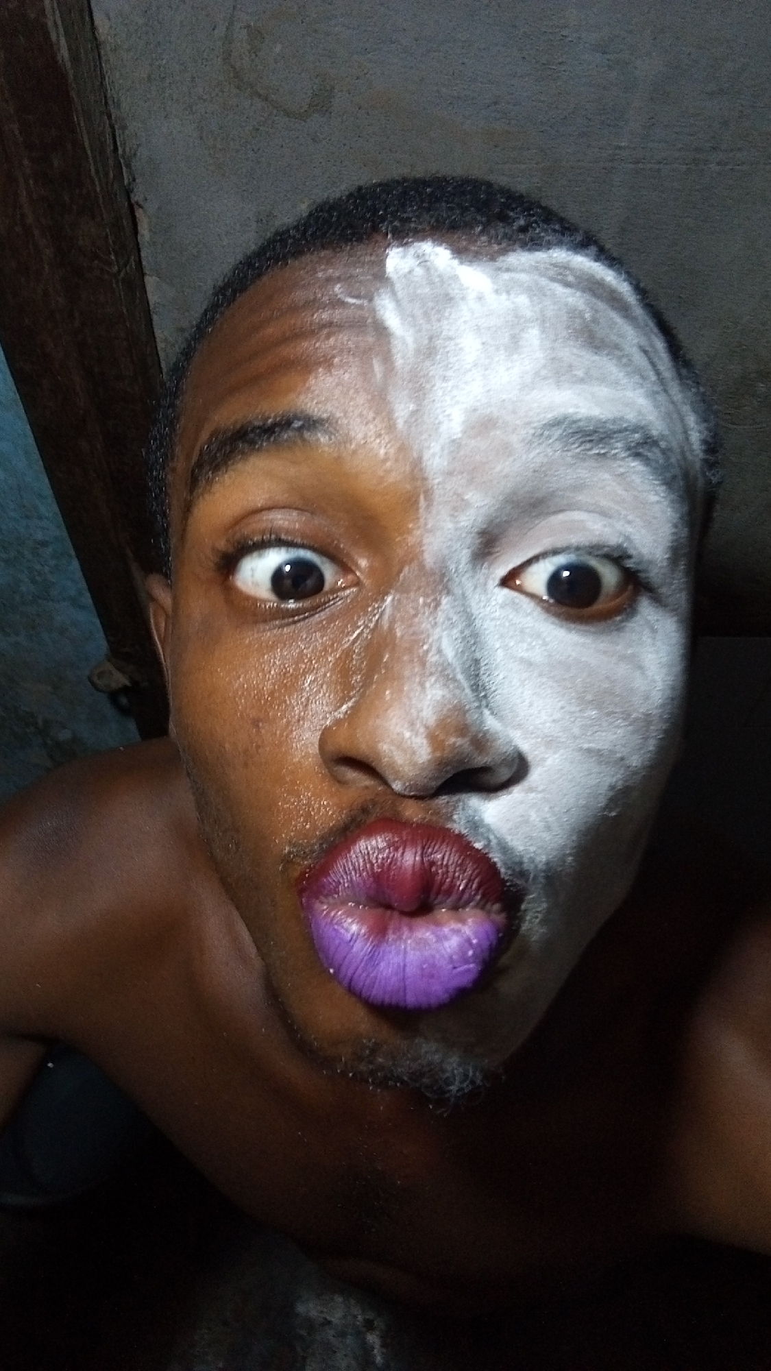 A close-up portrait features a person with dark skin, whose face is strikingly divided in appearance. The left side of the face (viewer's left) is uncovered, showing natural skin with some sheen, while the right side (viewer's right) is thickly coated in white powder or paint, creating a dramatic, two-tone effect. Both eyes are wide open, looking directly at the camera with a surprised or exaggerated expression. The person's lips are prominently puckered into a "kissing" pose and are painted a vibrant purple. Visible stubble can be seen on the chin and jawline. The background is dark and indistinct, appearing to be a rough, possibly concrete, wall or ceiling. A dark wooden beam is partially visible on the far left side of the frame. The scene is dimly lit, focusing attention on the person's expressive face and unique makeup. There is no visible text.