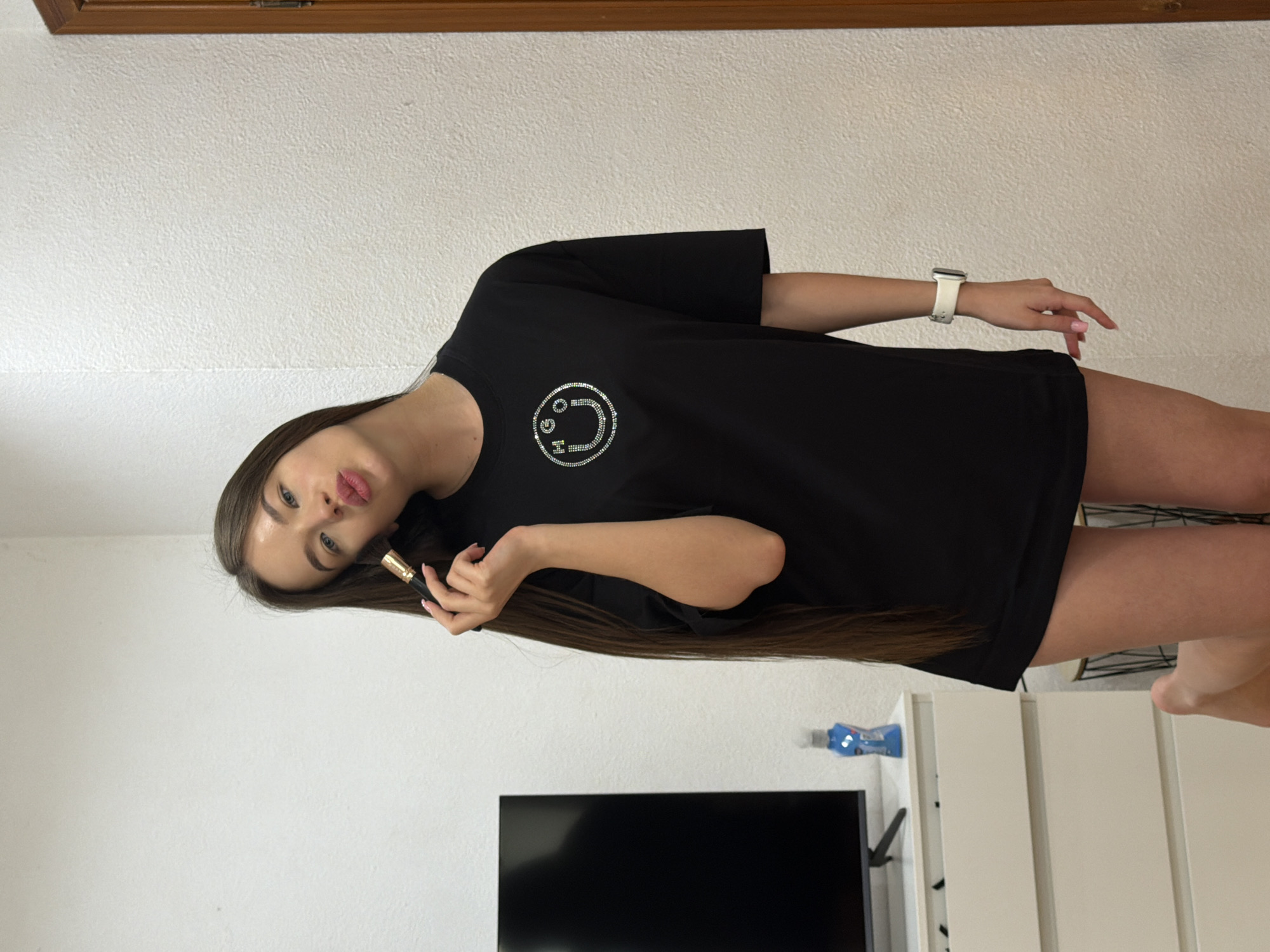 A woman is posing in a room. She is wearing a black t-shirt with a smiley face design. She is holding a makeup brush in her hand. The room contains a television, white cabinets, and a wooden trim. The city of Kovel, Ukraine, is not identifiable from the image.