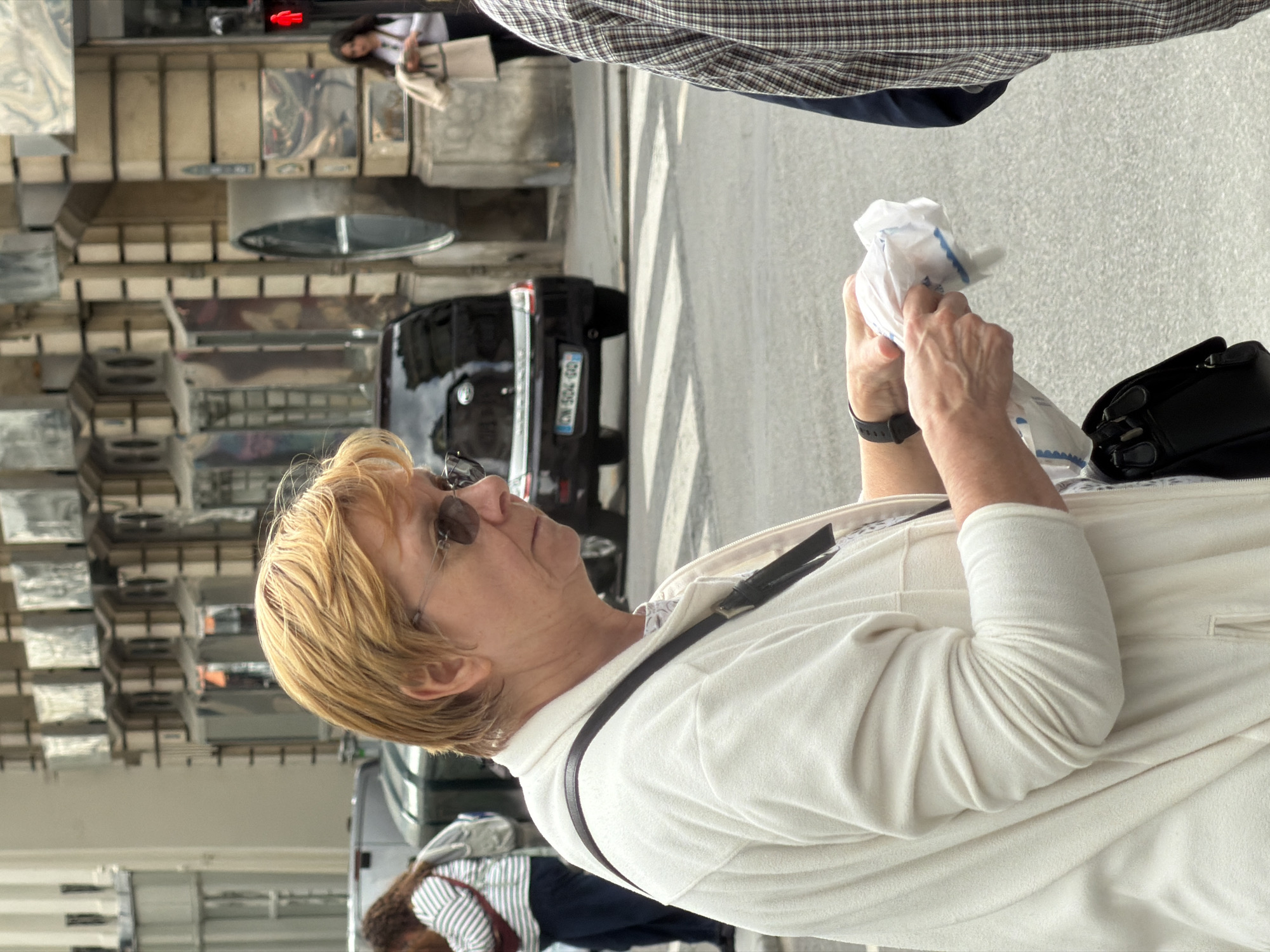 A medium shot of a fair-skinned woman, wearing sunglasses and a white jacket, holding a small white packet with a blue scalloped edge is displayed with a black purse next to her. A person wearing a striped shirt is situated near the bottom of the shot. Behind the woman, a black SUV with license plate "CN 504 GD" is visible on an urban street. Beyond the vehicle, a building with a unique, reflective, rectangular facade is evident. A crosswalk is marked on the pavement, and a pedestrian is discernible in the distance. Given the architectural style, reflective facade, and license plate format of the SUV, the photograph appears to be taken in Paris, France.