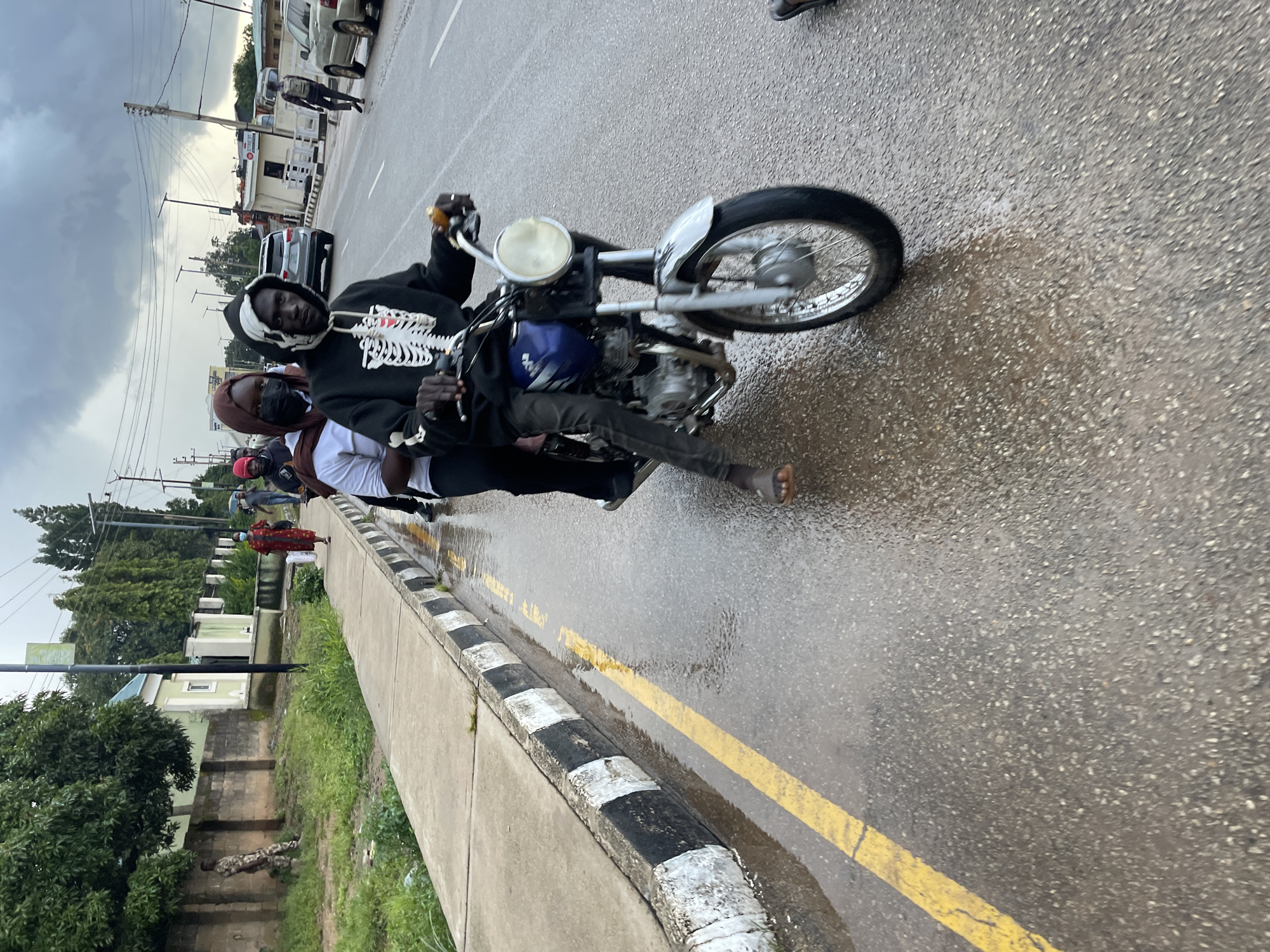 This image, captured in the city of Jos, Nigeria, depicts a scene on a wet asphalt road, likely after rainfall, as evidenced by the puddles and reflections. In the foreground, three people are riding a motorcycle. The driver, wearing a black hoodie with a white skeleton design, is in control, while two passengers are seated behind him. One passenger wears a headscarf and a black face mask, and the other is partially obscured. The motorcycle is blue and silver with a visible engine and front wheel.
To the left of the road, a concrete sidewalk with a black and white striped curb runs alongside a grassy area. Beyond the grass, a low wall and a building with a light green facade are visible. Further in the background, other vehicles, including a silver SUV and cars, are present on the road. The sky is overcast, contributing to the subdued lighting of the scene. People can also be seen walking on the sidewalk in the middle ground, one of whom is wearing a red dress. The overall atmosphere suggests a typical day in an urban environment, with the recent rain making the road surfaces slick.