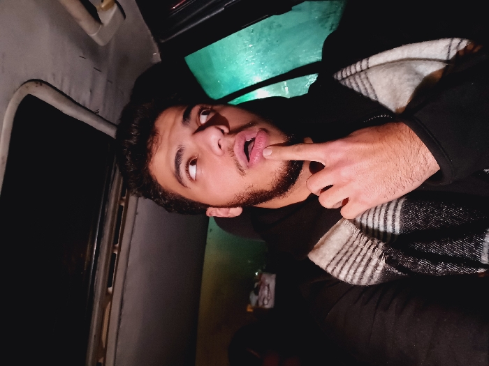 A young man with dark hair and a beard is facing upwards with his mouth open and finger on his lower lip. He is wearing a black jacket and a white and black striped scarf is draped around his shoulders. The background appears to be the interior of a vehicle. The walls and ceiling are light gray and there is a window visible on the left. There is also a glimpse of green light on the other side.