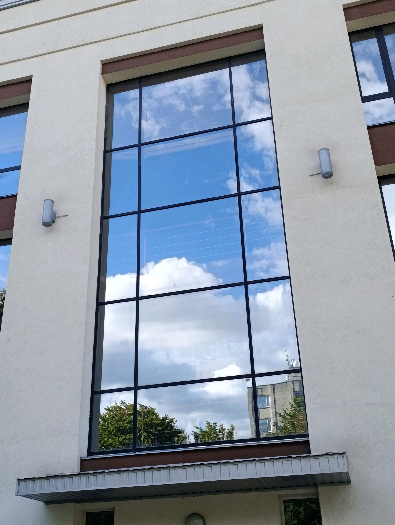 A low-angle outdoor medium shot captures the facade of a modern building with large, dark-framed windows reflecting a cloudy blue sky and greenery. The building's exterior is a light, textured material, likely stucco, with horizontal banding near the top. Two cylindrical, light-grey wall-mounted fixtures, likely outdoor lighting, are visible on the wall between the windows. A metal awning with a ribbed surface extends from the building below the main windows, and a dark window is visible beneath the awning. The scene is devoid of people. The image was taken in Cherkasy, Ukraine.