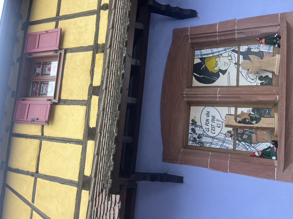 A two-story building facade in Kintzheim, France, is depicted from a low-angle perspective. The upper story exhibits a half-timbered construction with yellow infill panels and dark brown structural beams. A single rectangular window on this upper level features dark red wooden shutters; the left shutter is closed, while the right shutter is open, revealing a dark-framed window with white curtains patterned with red designs. Below the upper story, a terracotta-tiled roof with dark wooden supporting corbels extends over the lower section of the building. The lower story is painted a solid periwinkle blue. Centered on this blue wall is a large, rectangular window with a prominent reddish-brown stone frame. This window is divided into two panes. The left pane displays a comic-style illustration featuring a speech bubble with the French text "LE BON VIN C'EST PAR ICI !!" (The good wine is here!!) above a depiction of a potted plant with pink flowers. The right pane shows a comic-style illustration of a person with blonde hair and a dark hat, appearing to smile or wink. On the exterior sill of this lower window, two small figures resembling elves or gnomes, dressed in green and red, are seated. Between them are two wooden decorative cutouts: a stylized wooden Christmas tree with a yellow star on the left, and an unpainted wooden angel cutout on the right.