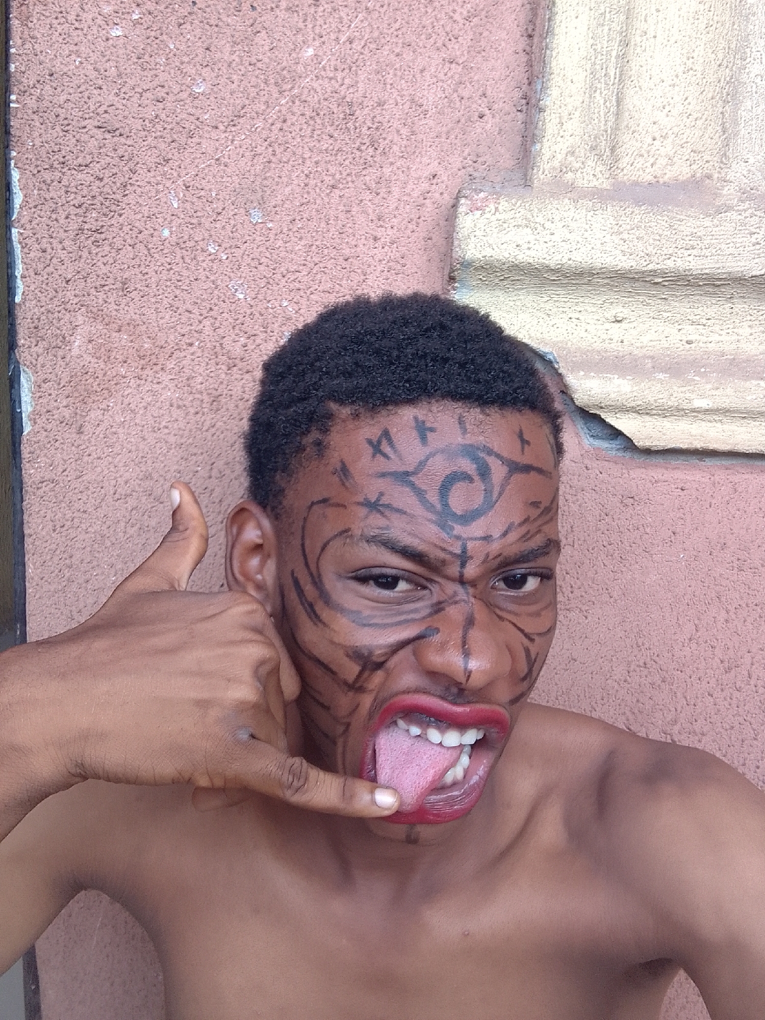 A young man with short, dark, curly hair is prominently featured, posing against a textured wall. His face is elaborately painted with intricate black designs, including a large, swirling symbol on his forehead, and bold lines extending around his eyes, cheeks, and chin, creating a tribal or character-like appearance. He is shirtless, revealing his bare shoulders and chest.
The man's expression is dynamic and energetic; his eyes are wide and focused, and his mouth is open in a wide grimace, revealing white teeth and a red tongue sticking out. His lips are colored with a vibrant red lipstick or paint. With his left hand, he makes a "shaka" or "call me" gesture, holding his thumb and pinky finger extended while the middle fingers are curled, positioned near his left cheek.
The background consists of a rough, light reddish-brown (or salmon-pink) textured wall. To the right, part of a lighter, off-white or cream-colored architectural structure with some detailing is visible. The lighting suggests it is daytime, with clear, bright conditions. The overall impression is one of a playful or fierce character portrayal, captured in a bold, expressive pose. There is no visible text in the image.